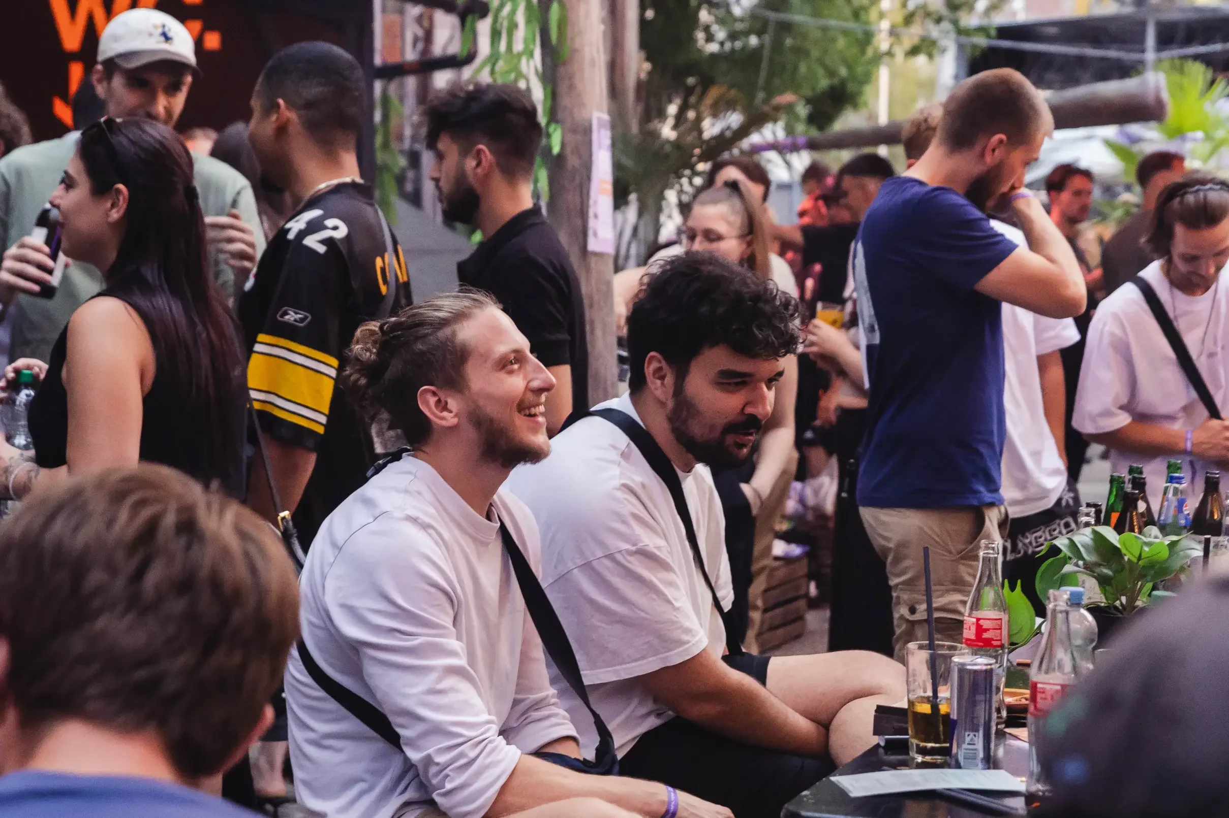 Menschen sitzen an einem langen Tisch bei einem Treffen oder einer Veranstaltung, einige unterhalten sich, andere lachen und trinken Getränke, im Hintergrund grüne Bäume und einige Hütten.