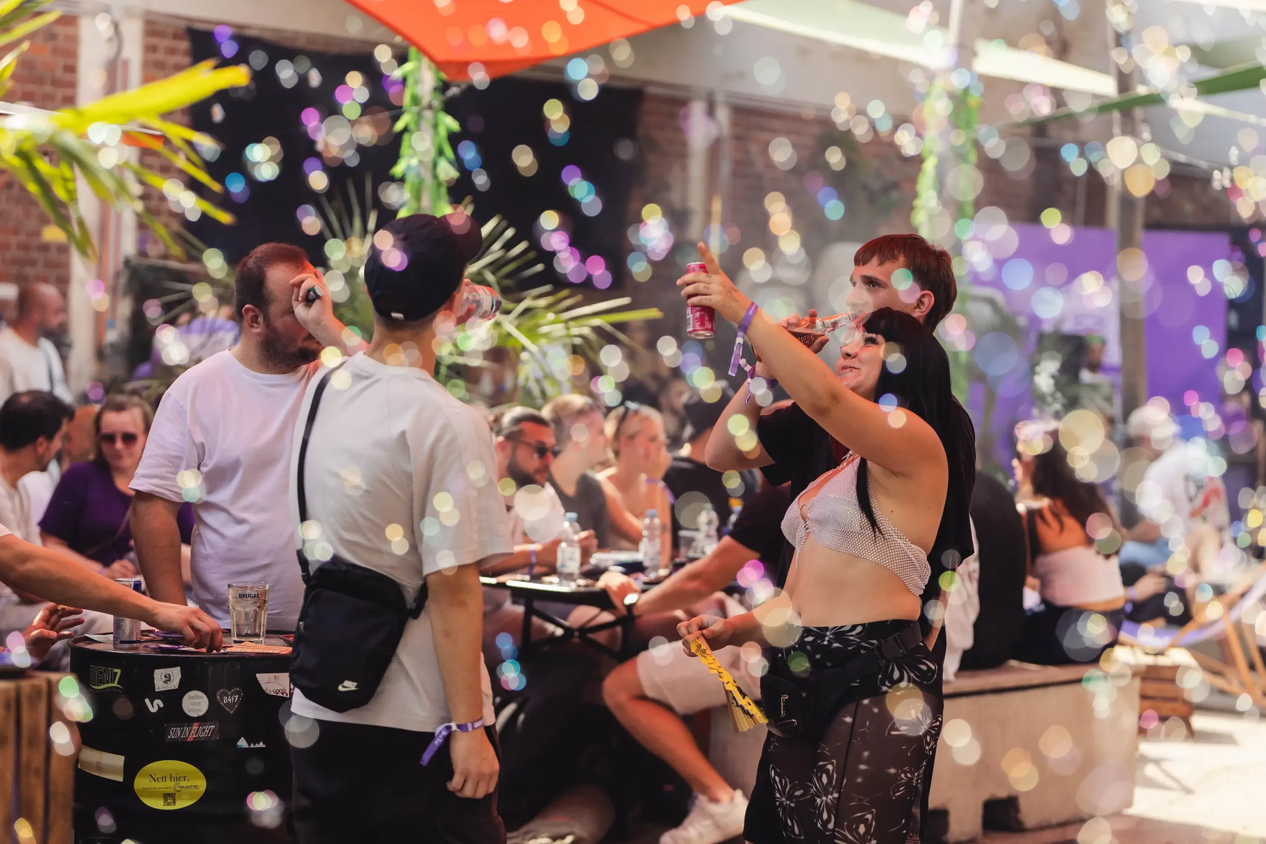Menschen feiern auf einer Party im Freien bei Nacht mit bunten Lichtern und Dekorationen, einige trinken und unterhalten sich.