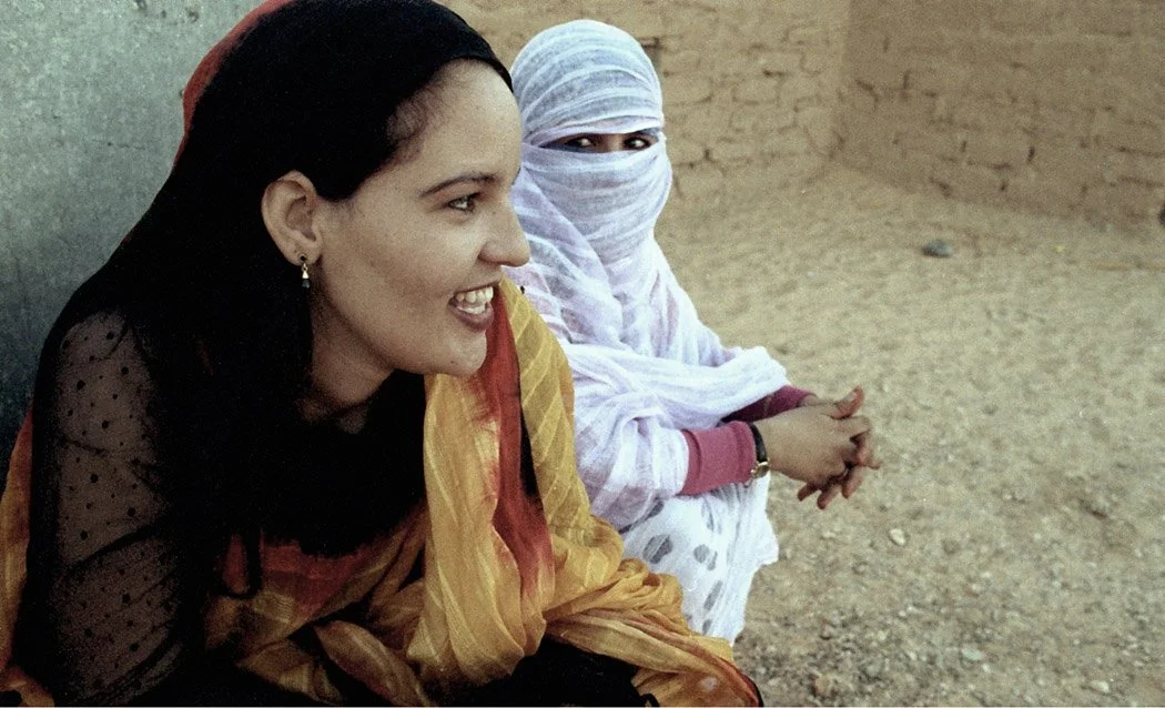 Two women sitting on the ground outdoors, one smiling and wearing a colorful traditional outfit, the other covered with a white headscarf and face covering.