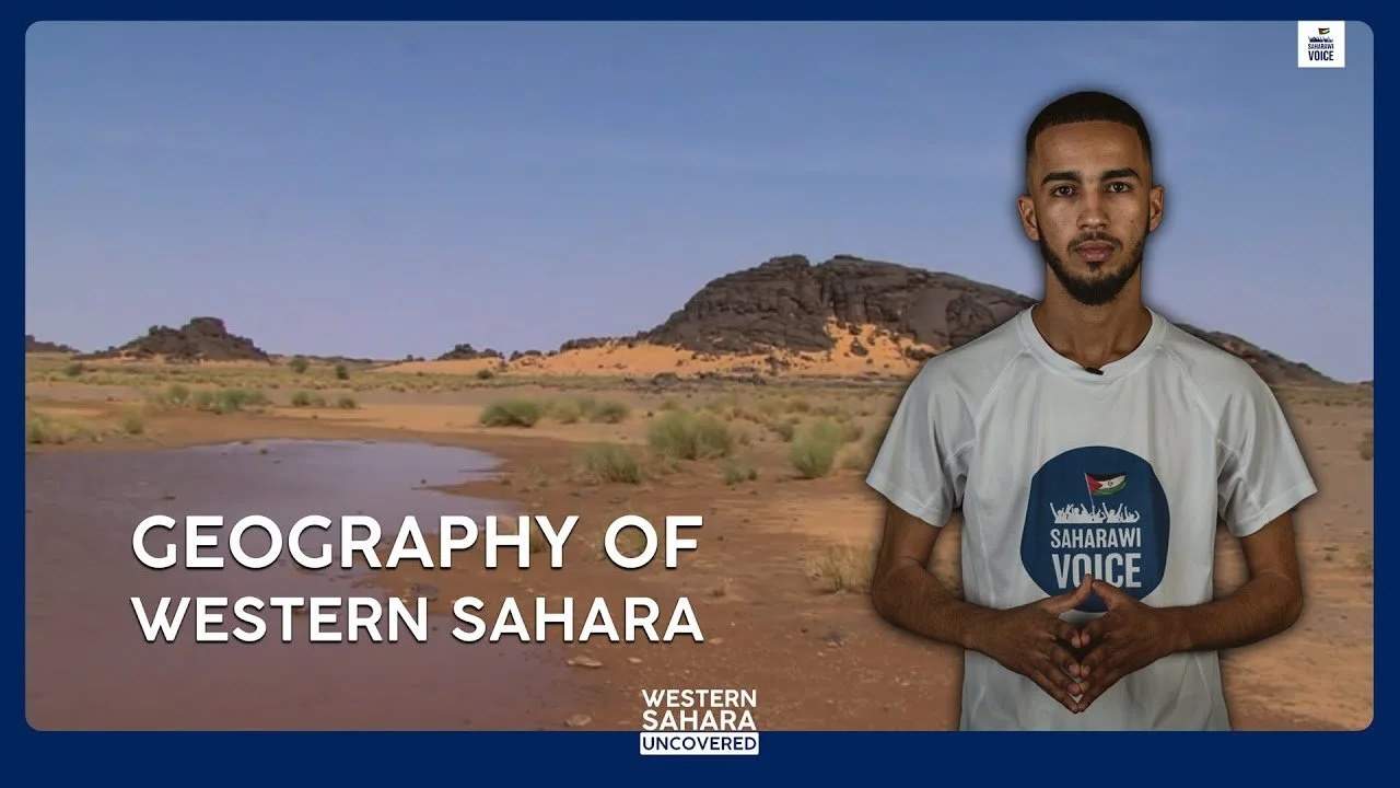 A man standing in front of a desert landscape with rocky formations and sparse vegetation, wearing a white t-shirt with the logo ‘Saharawi Voice’, holding his hands together, with text overlay reading ‘Geography of Western Sahara’.