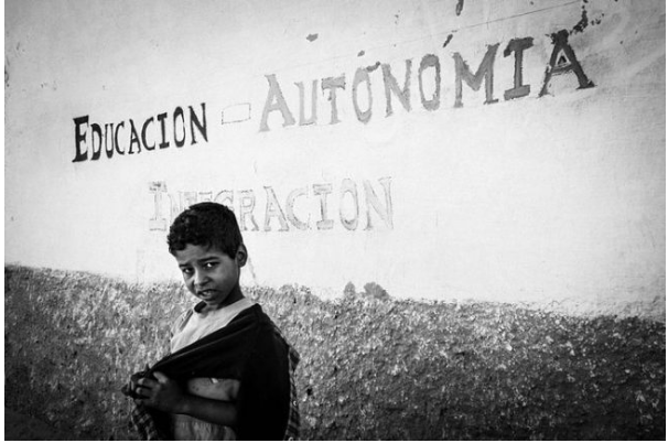 A young boy standing in front of a wall with painted words in Spanish, including 'Educacion,' 'Autonomia,' and 'Dignidad.'