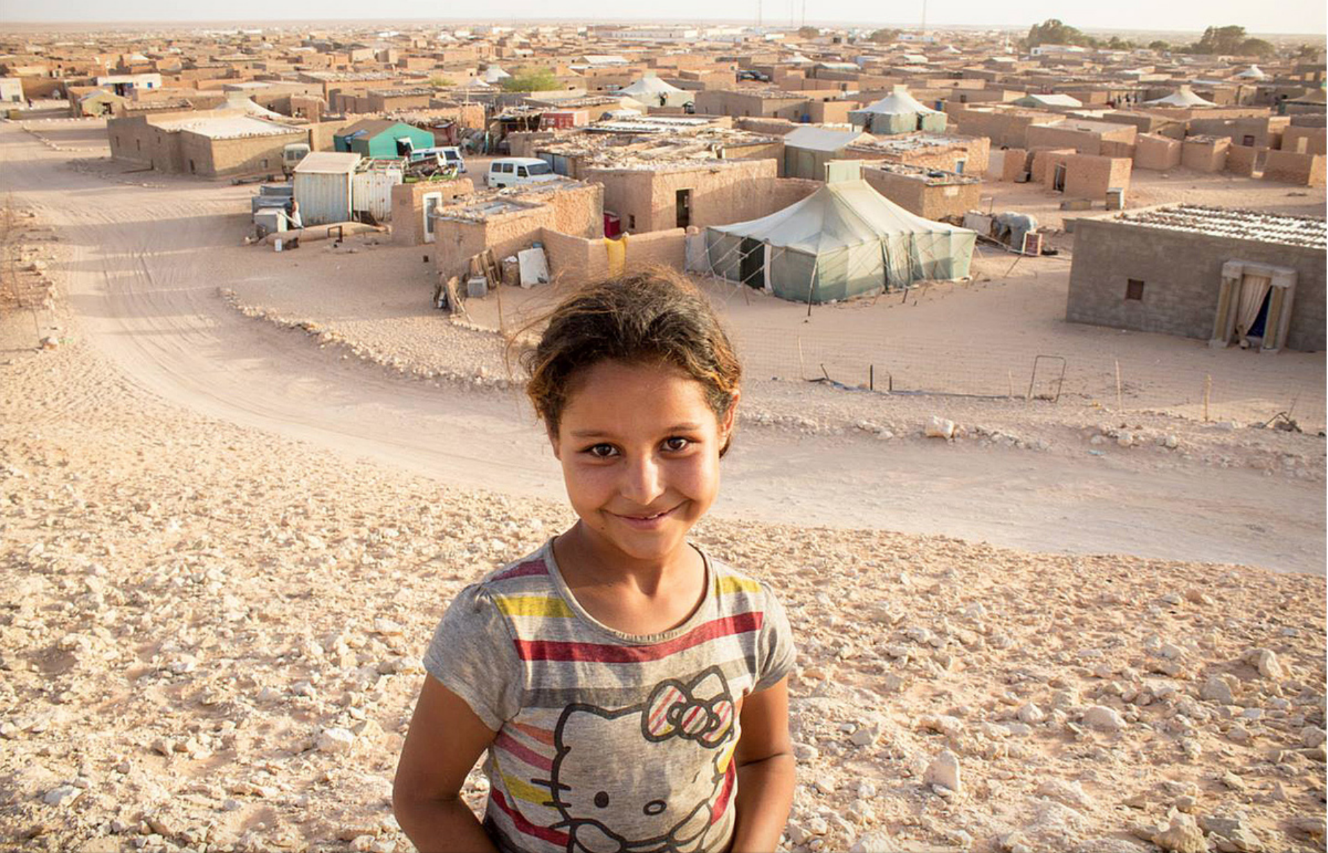 A young girl standing on a rocky hill in a desert village with adobe buildings and tents in the background.