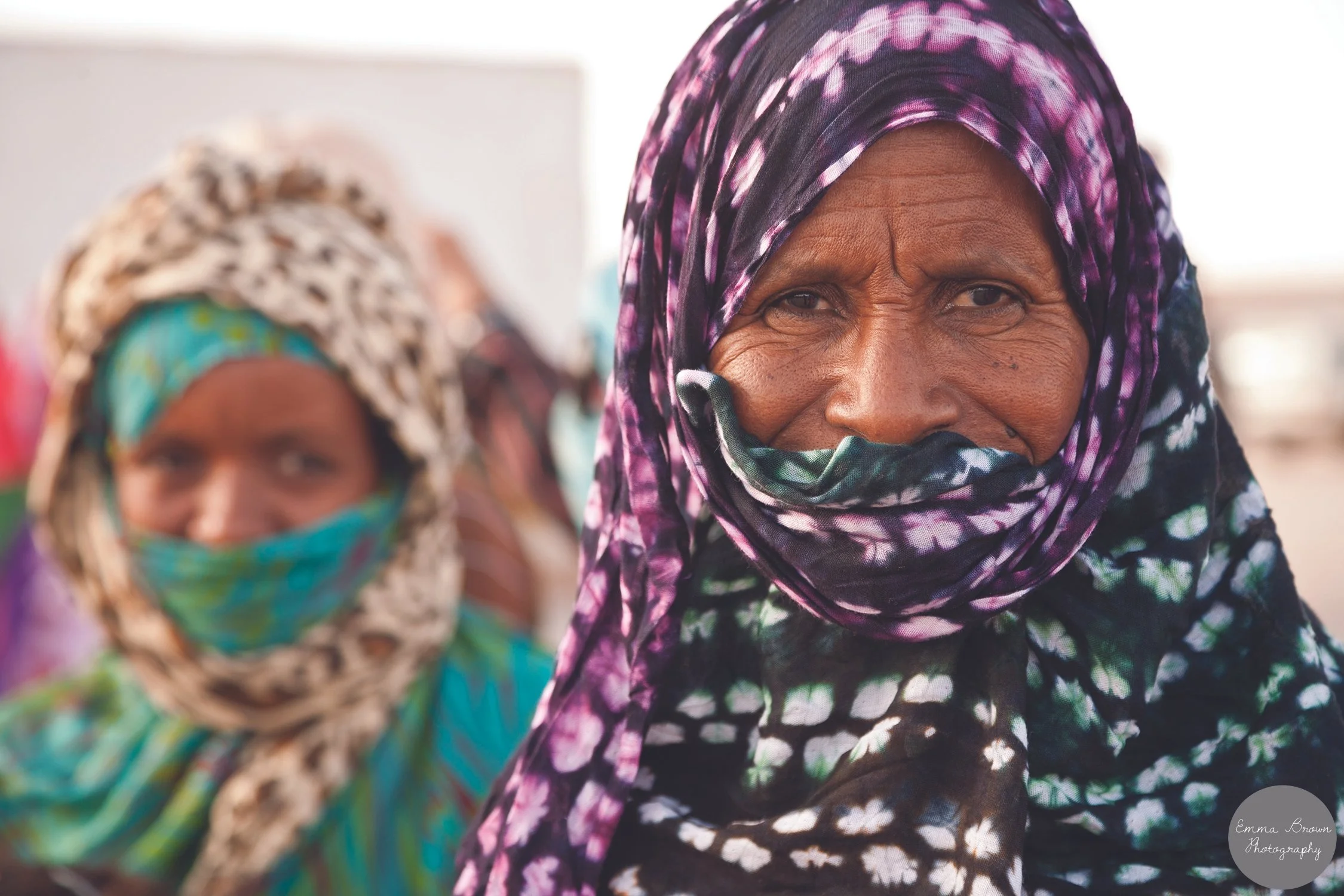 Close-up of an older woman wearing a purple and black tie-dye headscarf, with another woman wearing a patterned headscarf and face mask blurred in the background.