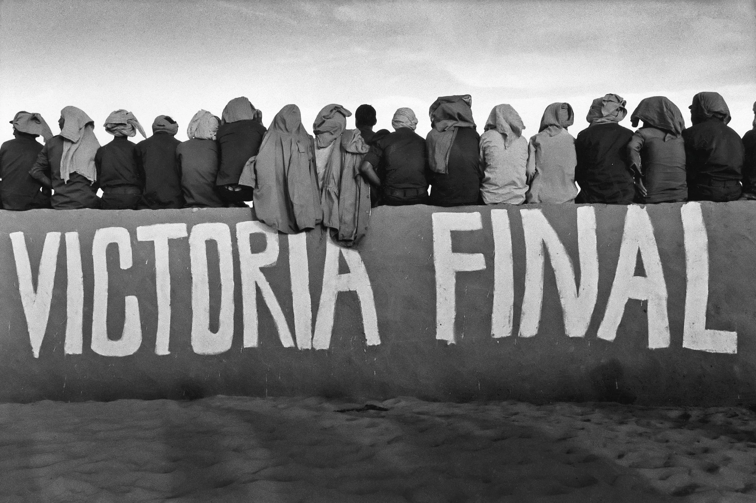 A group of people sitting on the edge of a structure with a large sign that reads 'VICTORIA FINAL'. The individuals are wearing head coverings and are shown from behind.