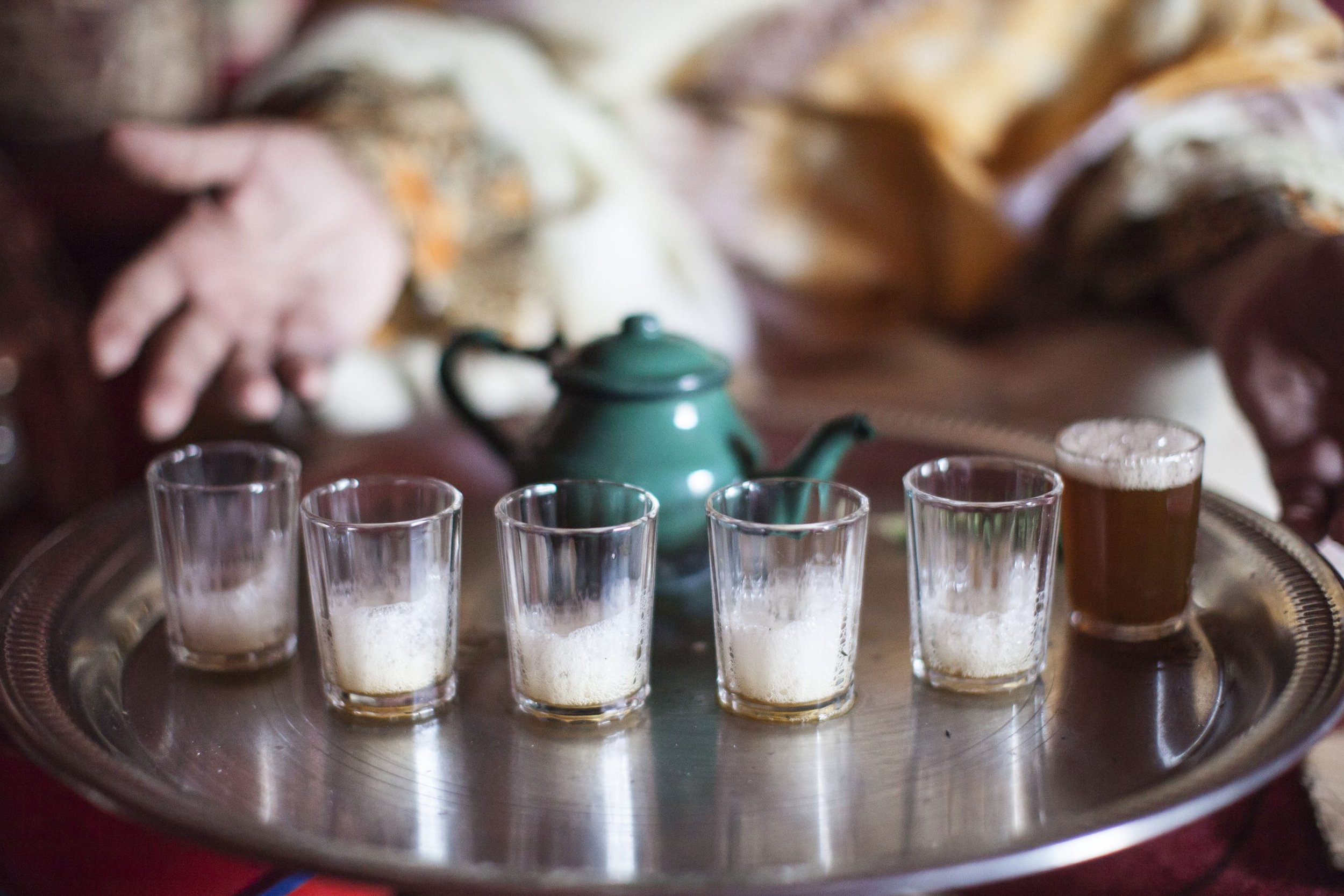 Five shot glasses filled with a foamy beverage on a silver tray, with a green teapot in the background and a person's hand reaching toward the tray.