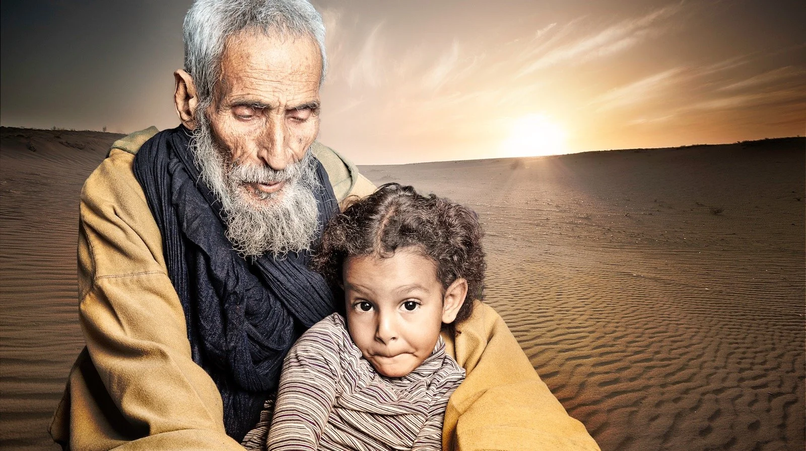 An elderly man with a gray beard and a young girl with curly hair, both sitting together in a desert at sunset.