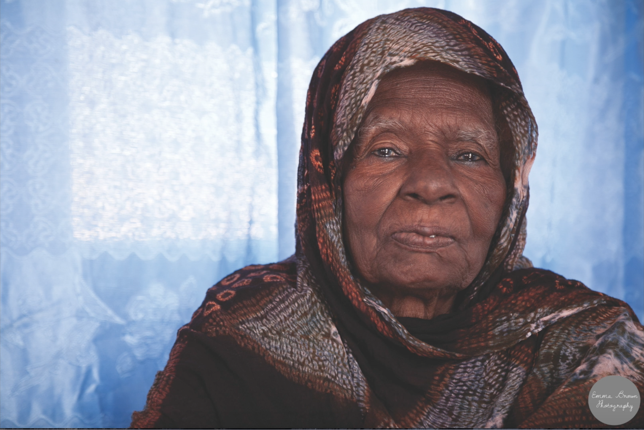 An elderly woman wearing a patterned headscarf and dark clothing standing in front of a light blue, lace curtain.