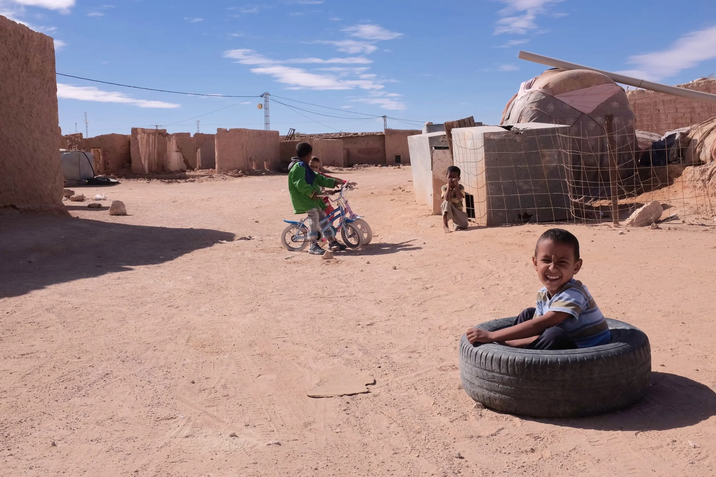 Children playing in a desert-like village, one sitting in a tire, others riding bikes, with mud buildings and a large blue sky in the background.