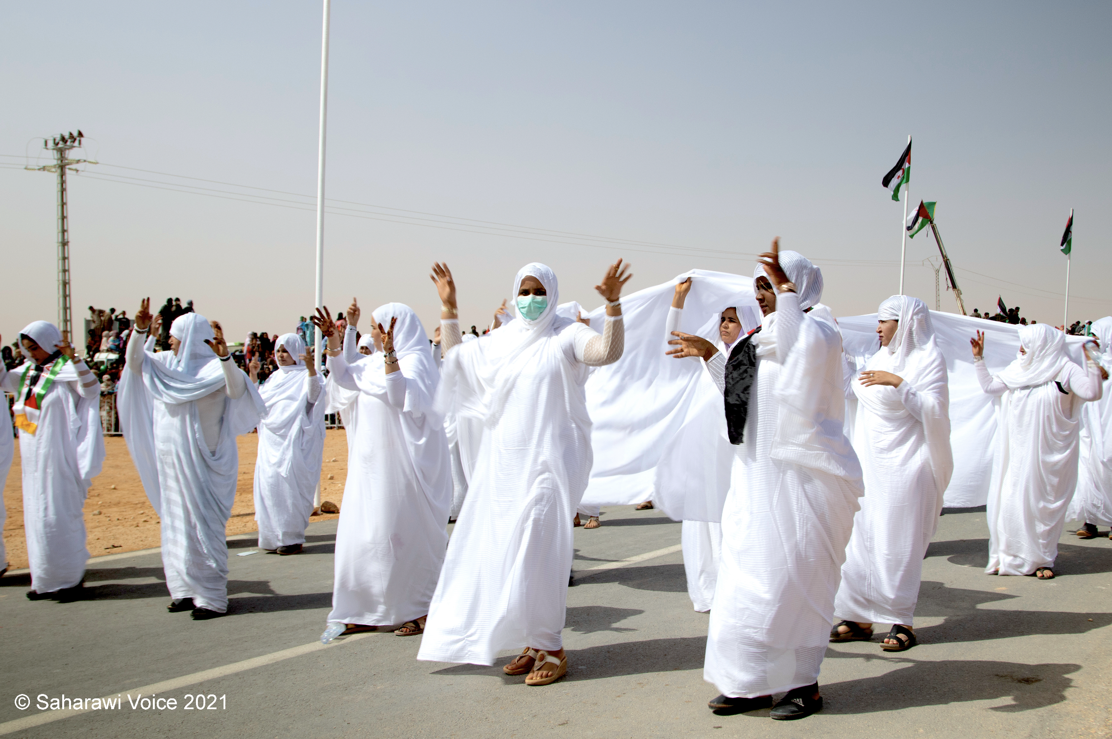 A group of women dressed in white traditional attire participating in a parade or celebration on a sunny day. Some women are wearing face masks. In the background, there are flags and a crowd of spectators.