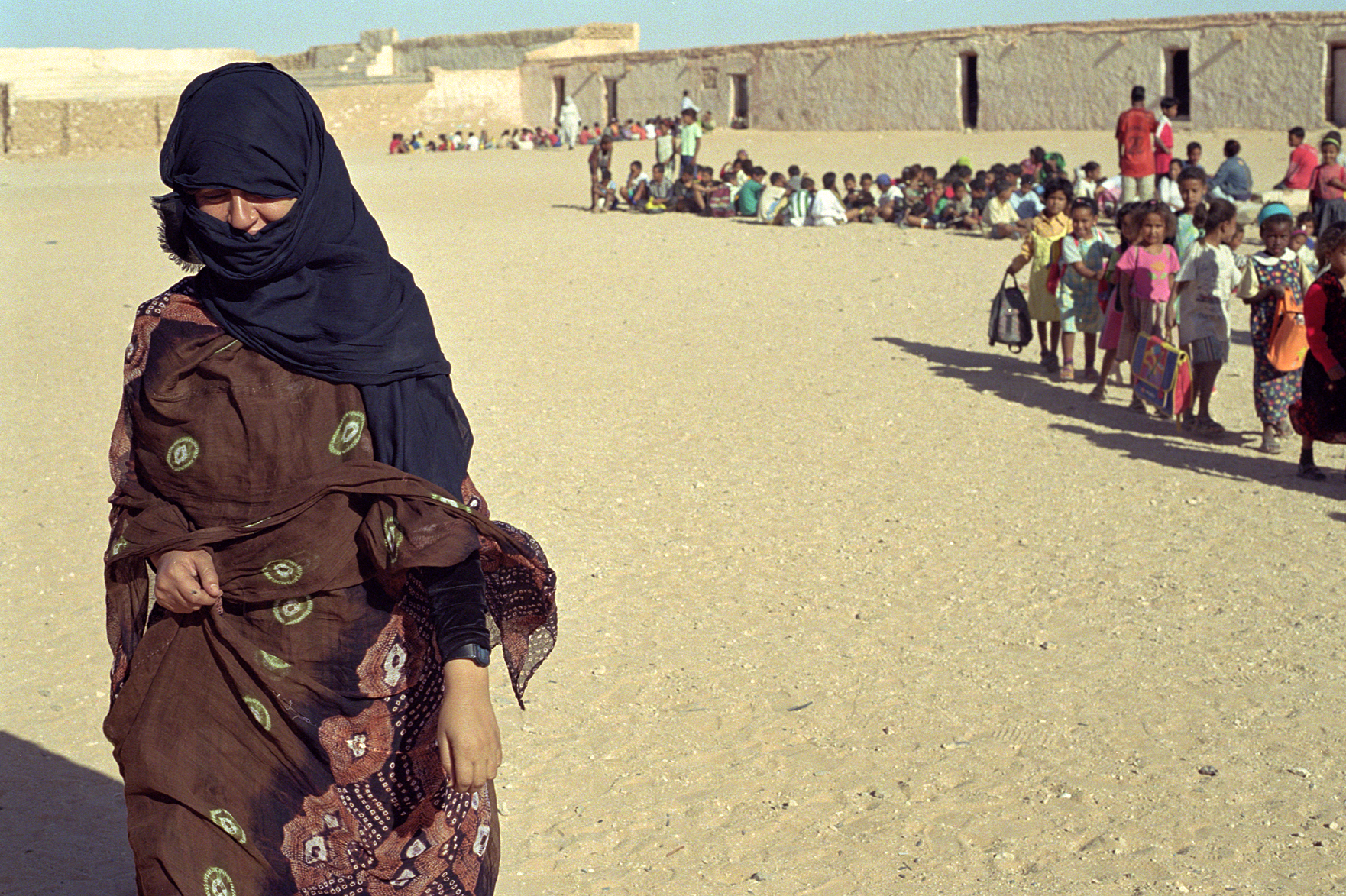 A woman in traditional attire, with a headscarf covering her face, walks past a long line of children with backpacks in a desert setting, near a building with small windows.
