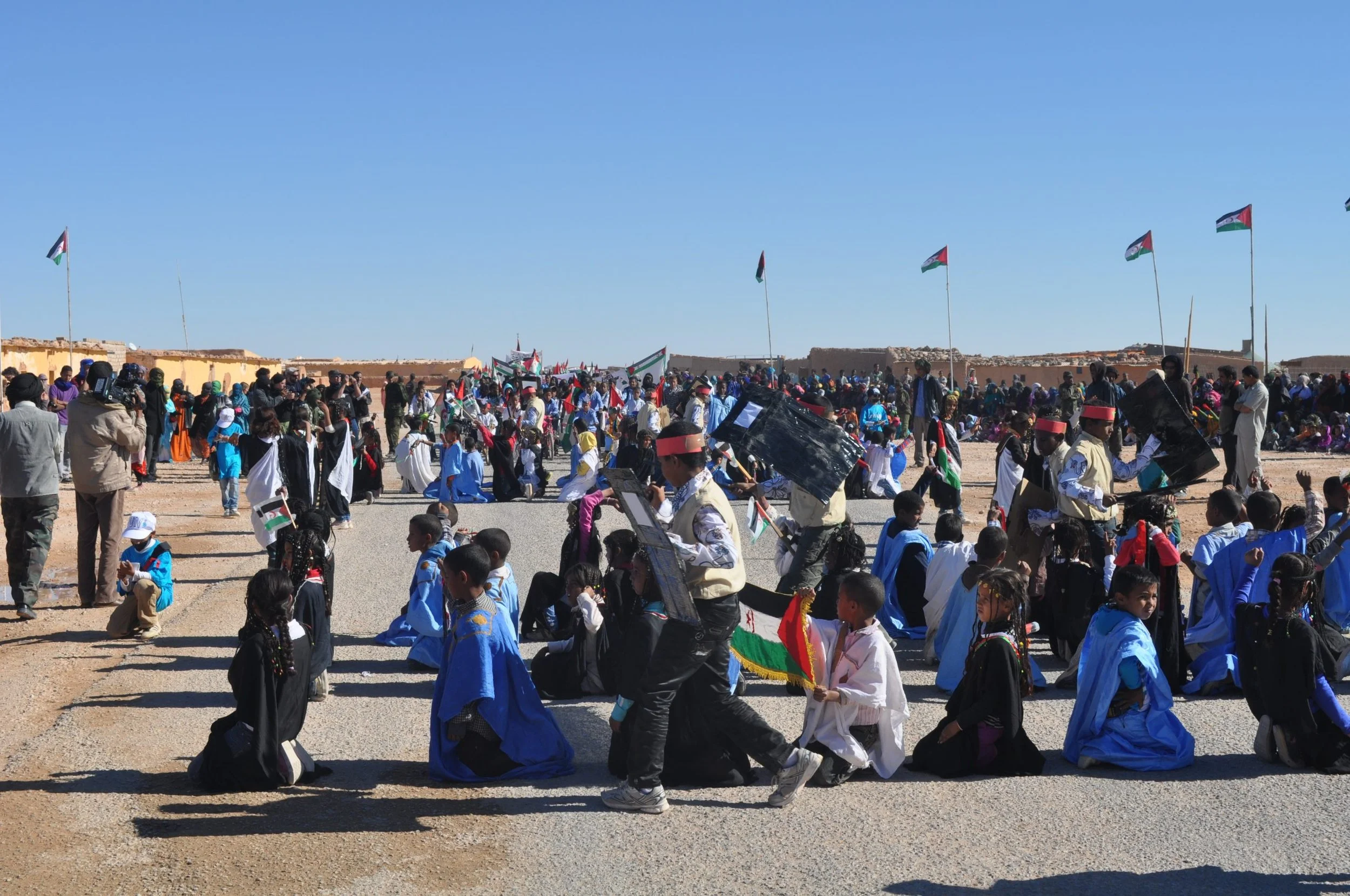 Large outdoor gathering with children and adults participating in a cultural event under a clear blue sky, with many flags and some onlookers.