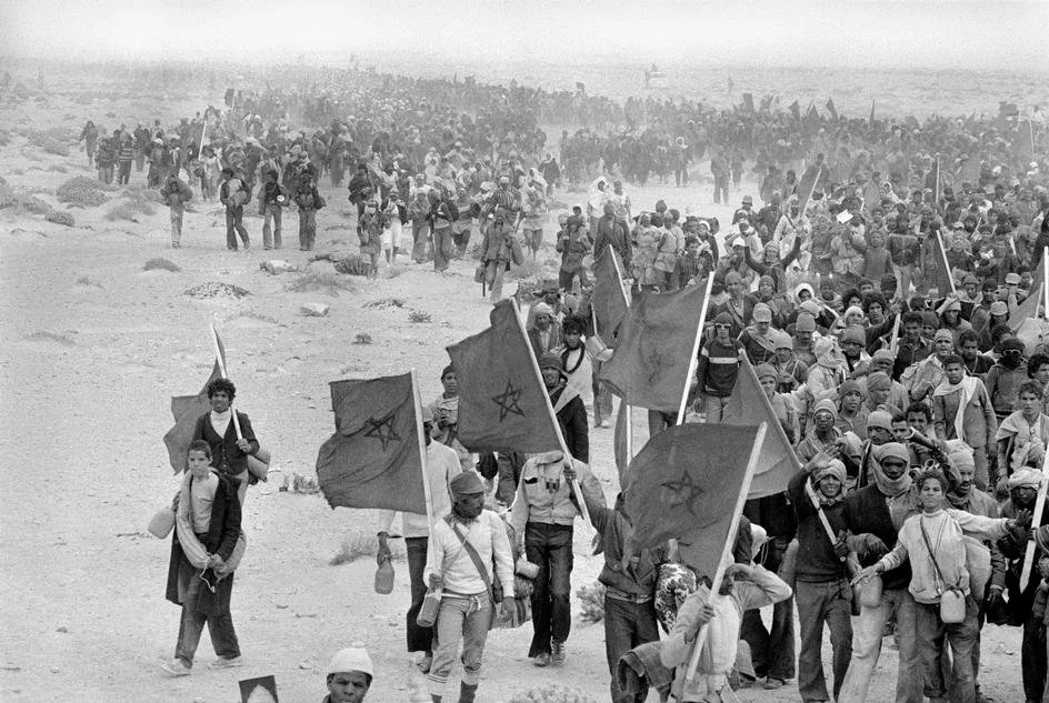 Large group of people marching with Moroccan flags on a beach or sandy area, some carrying backpacks and bags, in black and white.