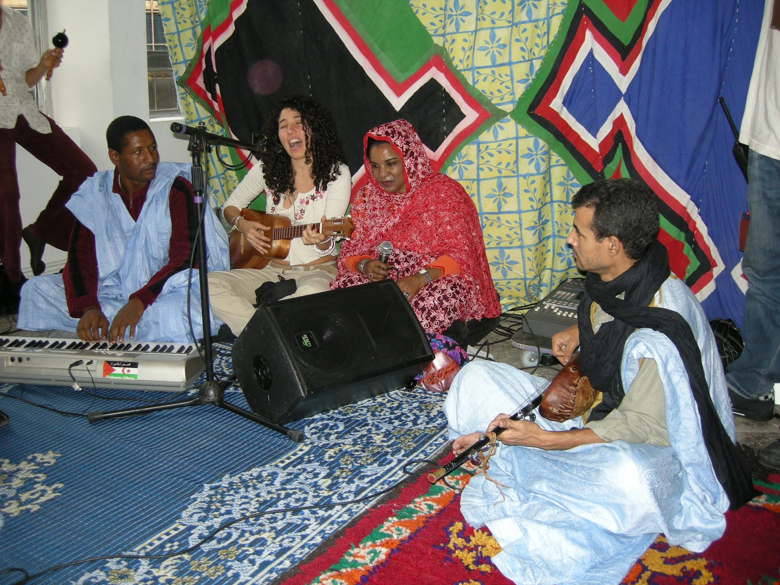 Group of people performing music together, sitting on the floor with musical instruments, colorful backdrop behind them.