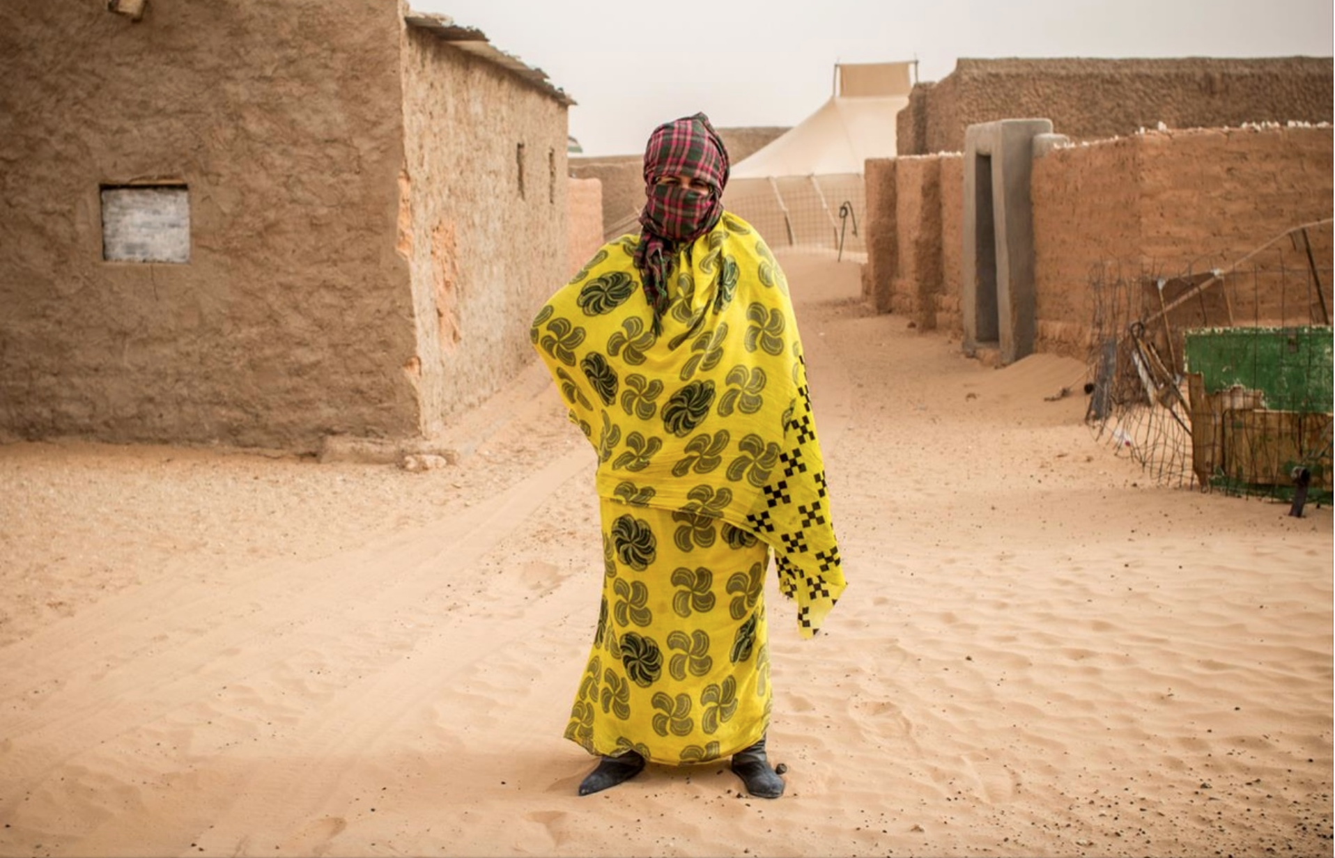 A woman wearing a yellow dress with black and green patterns, wrapped in the same fabric, standing on a sandy street in a desert village. She has her face covered with a scarf and is wearing a plaid headscarf.