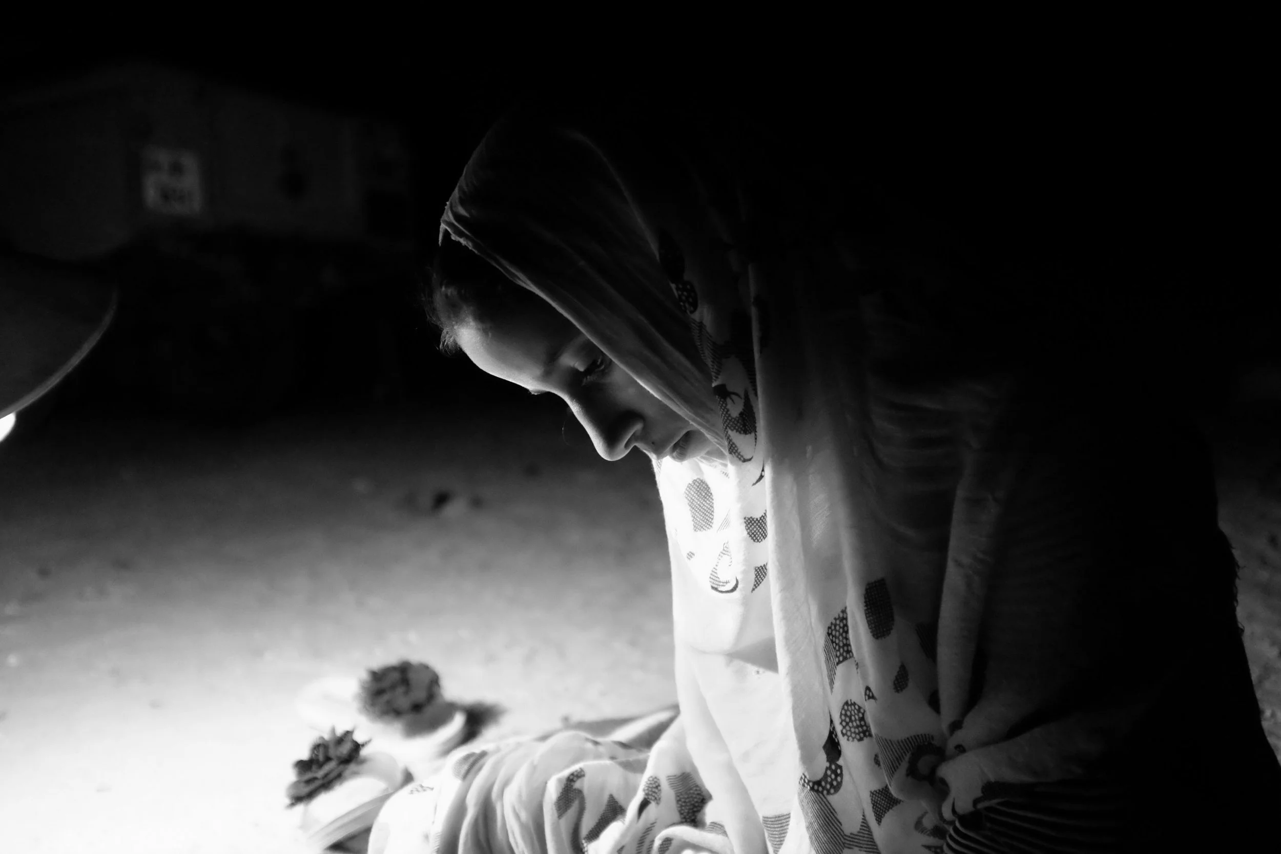 A young woman wearing a headscarf appears to be sitting or crouching on the ground, looking down with a focused or contemplative expression. The photo is in black and white, showing her profile and some objects nearby.
