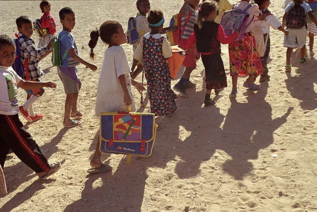 Group of children walking on a sandy beach, carrying backpacks and a school bag, with their shadows cast on the ground.