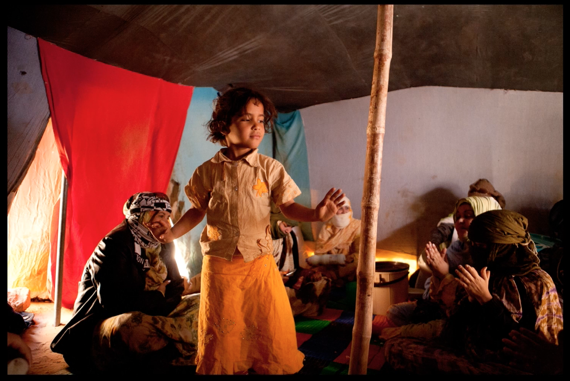 A young girl stands inside a tent, with several women sitting on the floor around her. The women are dressed in traditional clothing and head coverings, and are engaged in prayer or reflection. The tent has colorful fabric walls, including red and blue sections, and the scene is lit by warm, soft lighting.