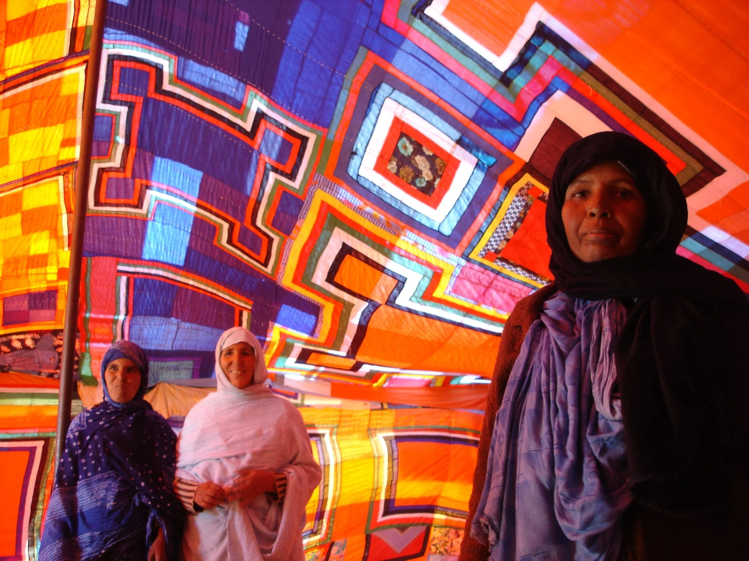 Three women inside a colorful geometric textile tent, with intricate patterns and vibrant colors.