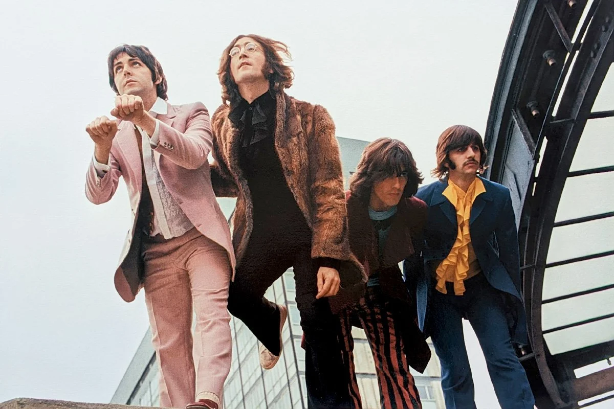 The Beatles in their iconic 'Sgt. Pepper's Lonely Hearts Club Band' era outfits, walking on a bridge during daytime.