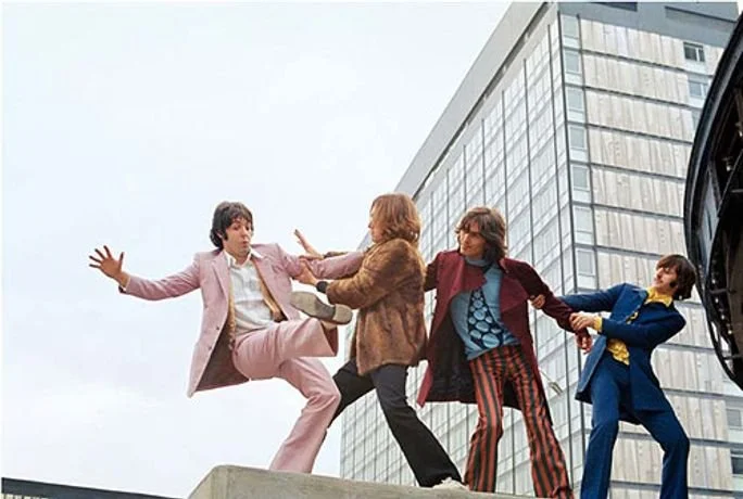 The Beatles dressed in 1970s attire appear to be fighting on a city rooftop with a modern glass building in the background.