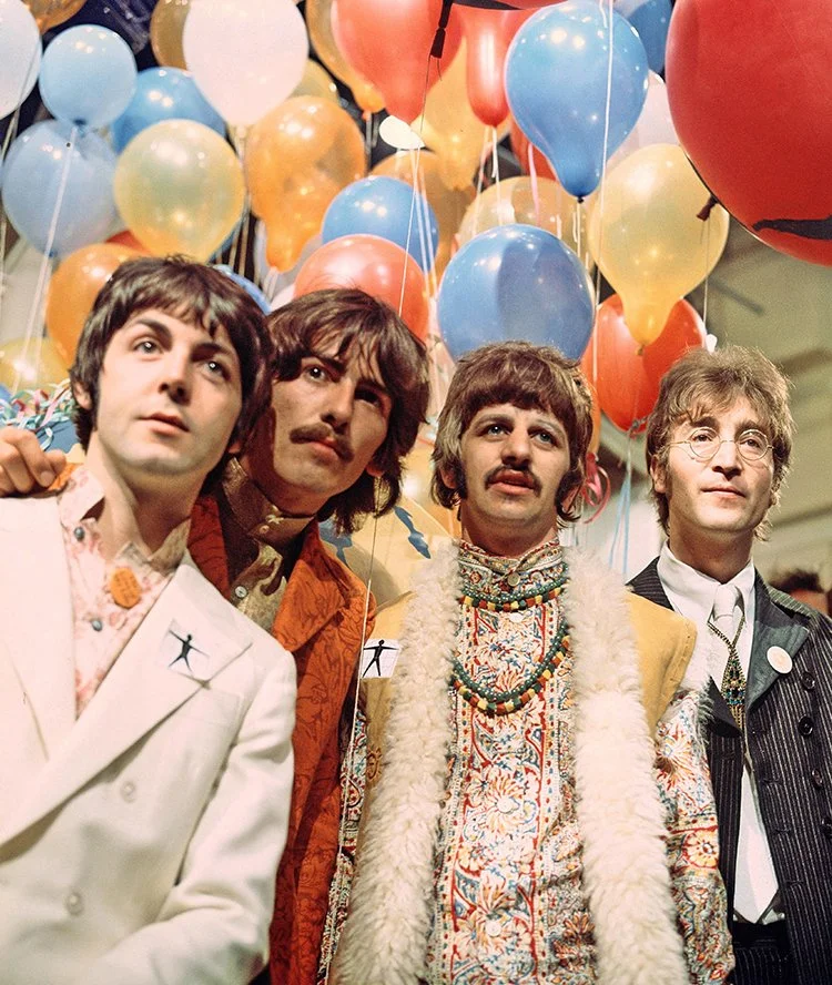 The Beatles dressed in 1960s fashion standing under a ceiling filled with colorful balloons, likely at a celebration or event.