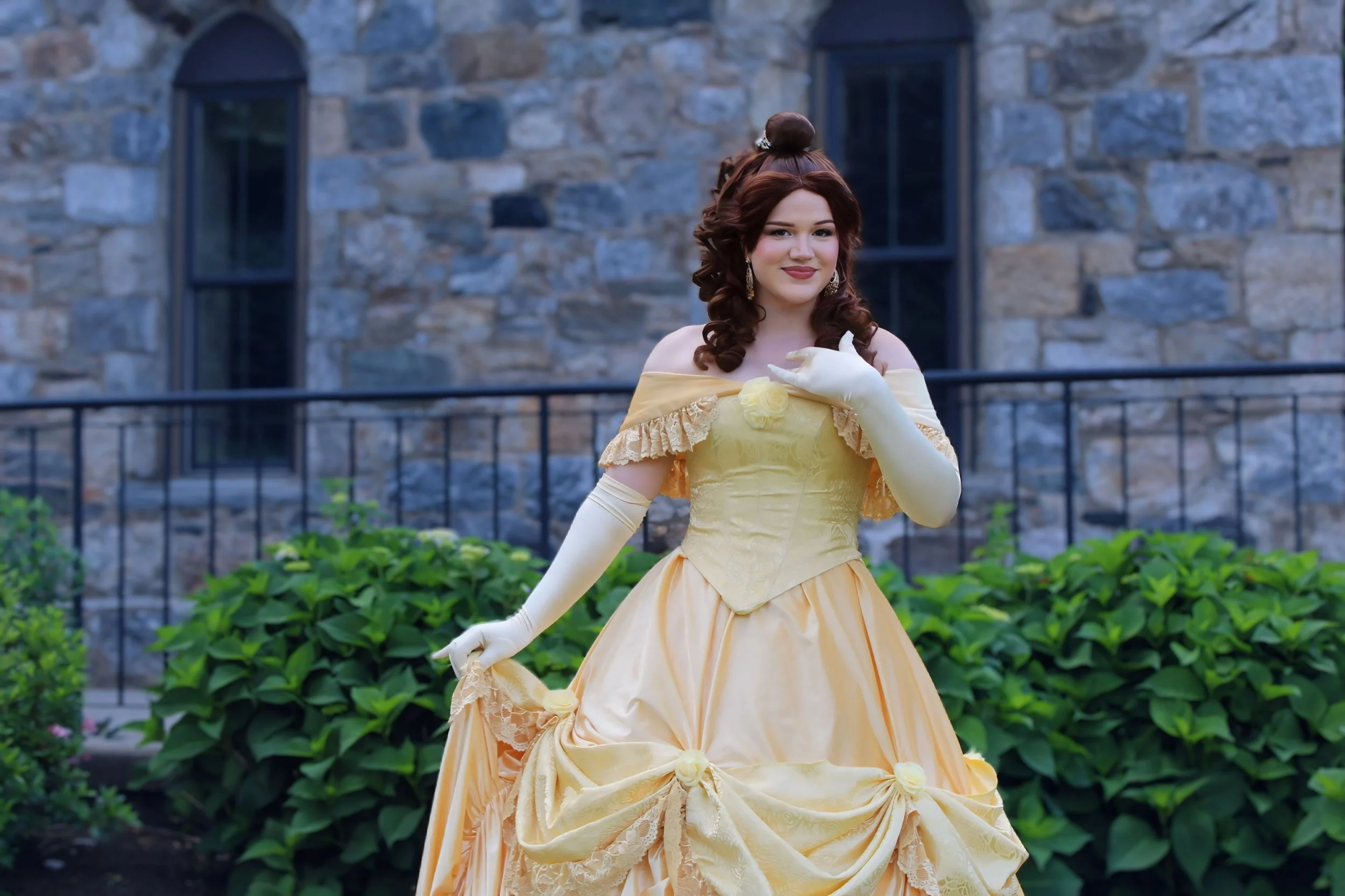 A woman dressed as Belle from Beauty and the Beast, smiling, wearing a yellow gown with gloves, with pink and pastel-colored paper butterflies and balloons in the background.