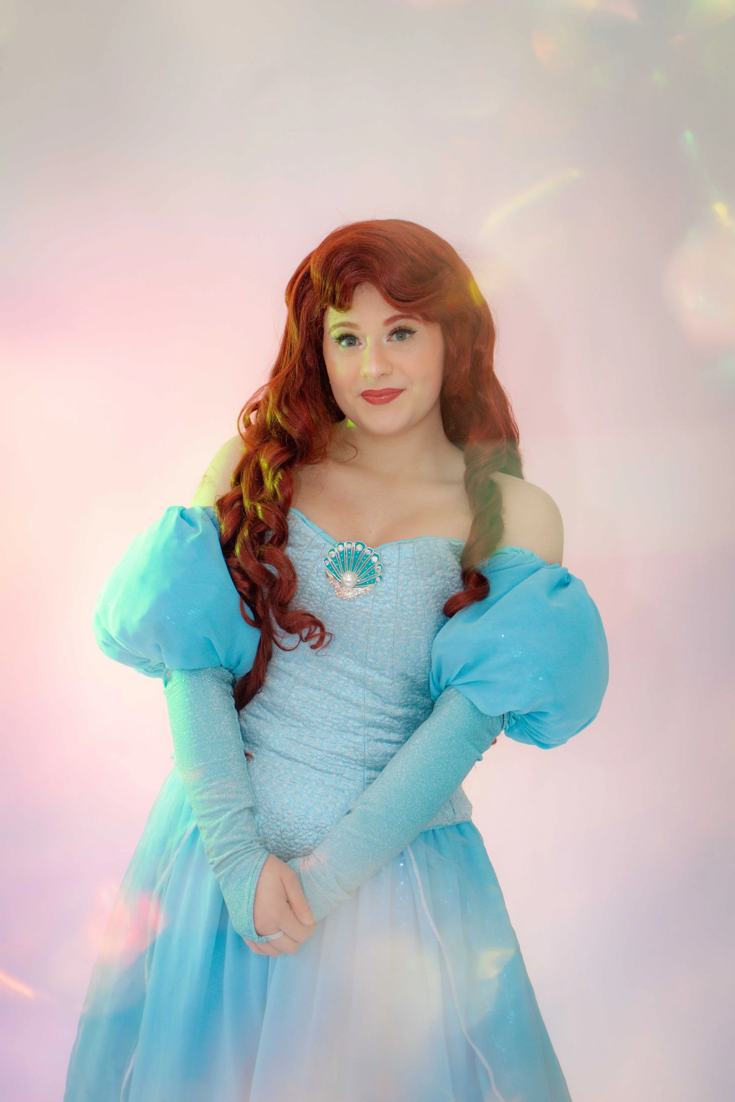 A woman dressed as Ariel from The Little Mermaid, with long red hair and a blue dress, standing in front of green foliage.