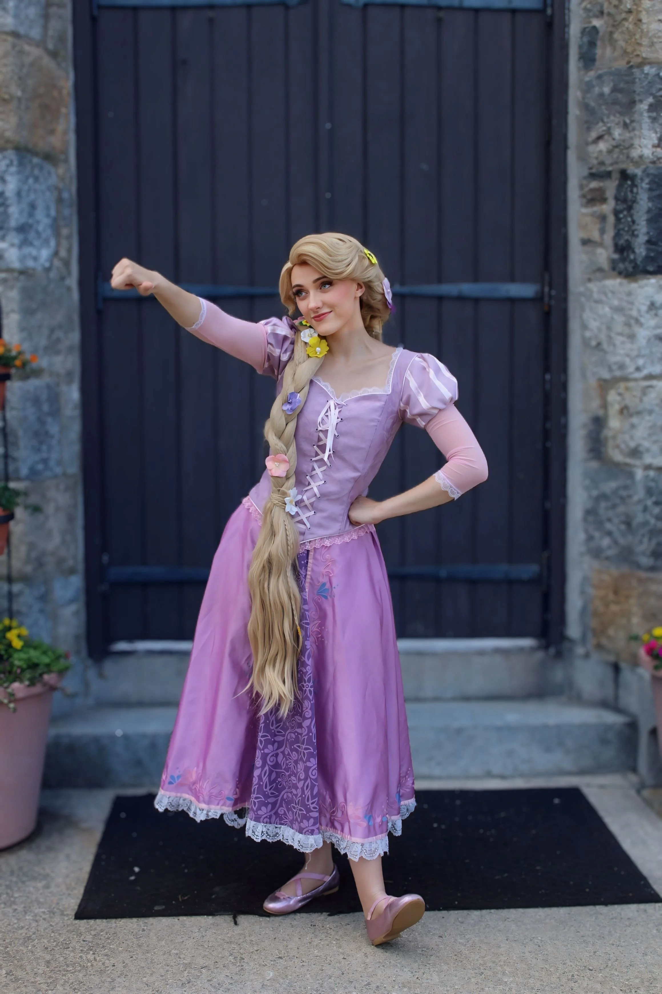Young woman dressed as Rapunzel from fairy tale, wearing pink dress with lace trim and a long braided blonde wig, standing against blue background, smiling and posing.