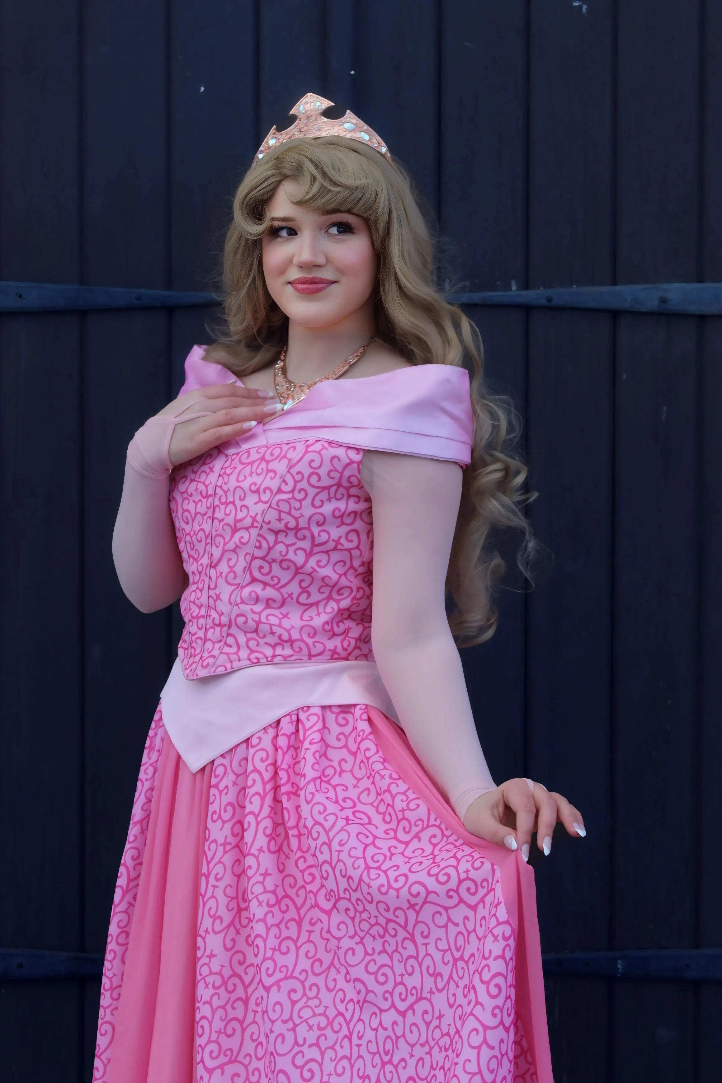 A woman dressed as a princess in a pink gown, wearing a tiara and necklace, standing in front of a dark wooden background.