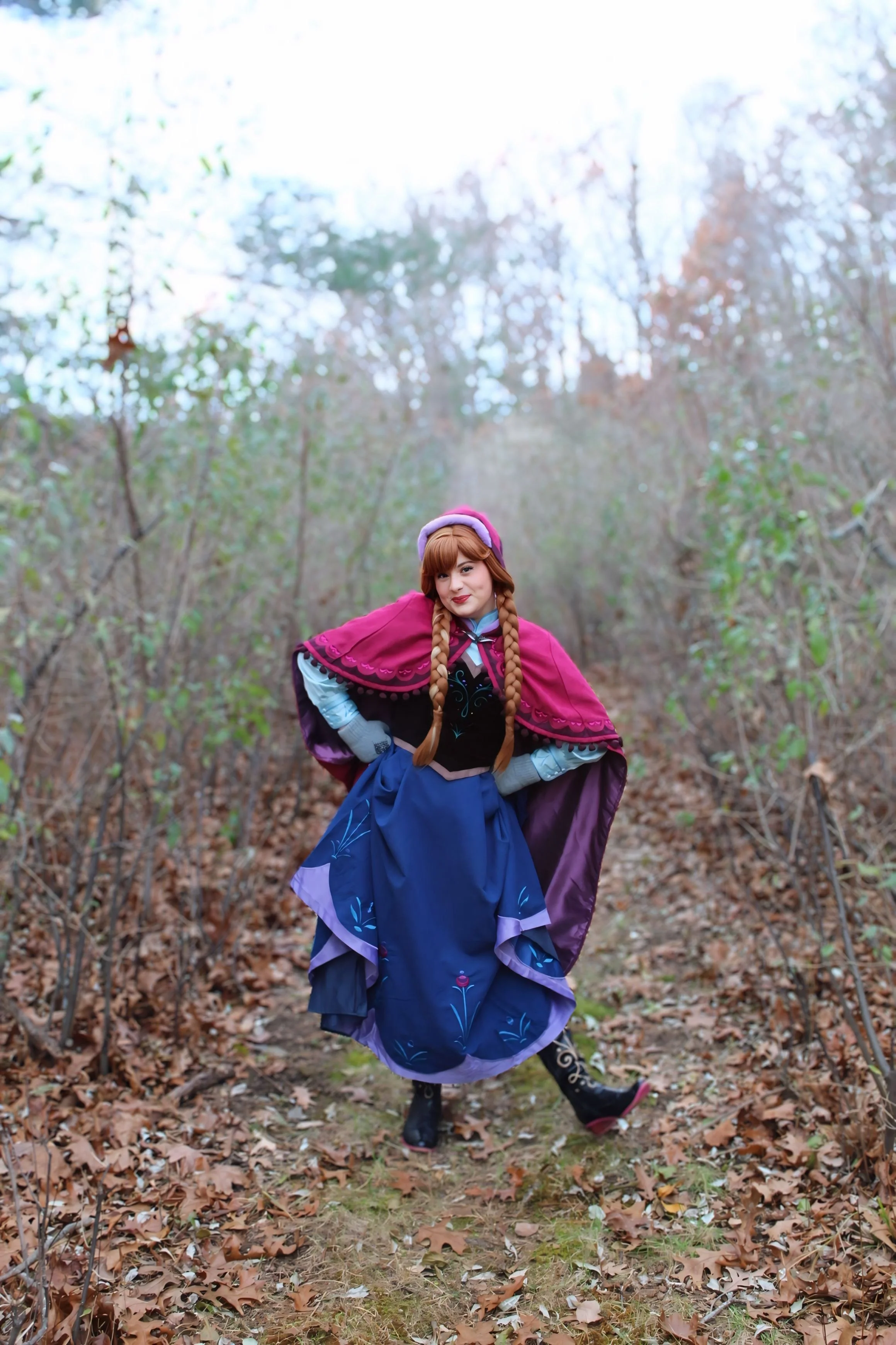 A woman dressed as Princess Anna from Disney's Frozen, with braided hairstyle, wearing a black and green bodice with embroidered designs, in an outdoor setting with greenery and trees in the background.