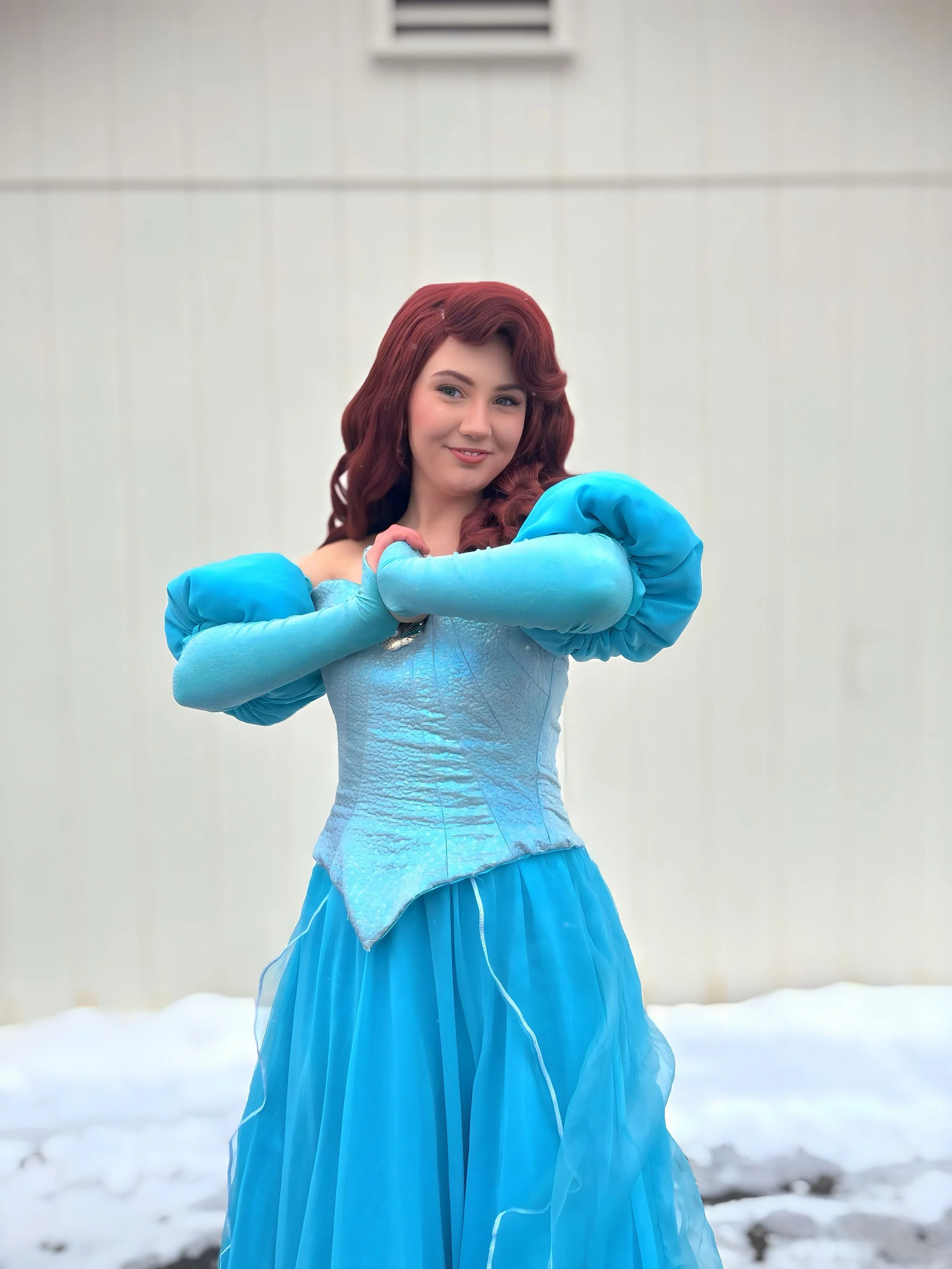 A woman dressed as Ariel from The Little Mermaid, with long red hair and a blue dress, standing in front of green foliage.