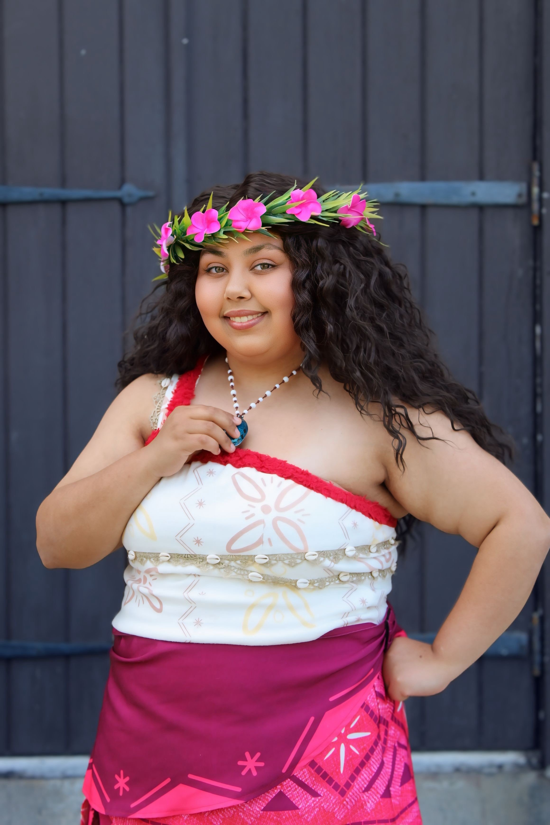 Woman wearing traditional Polynesian attire with a floral crown, colorful skirt, and jewelry, standing in a decorated room.