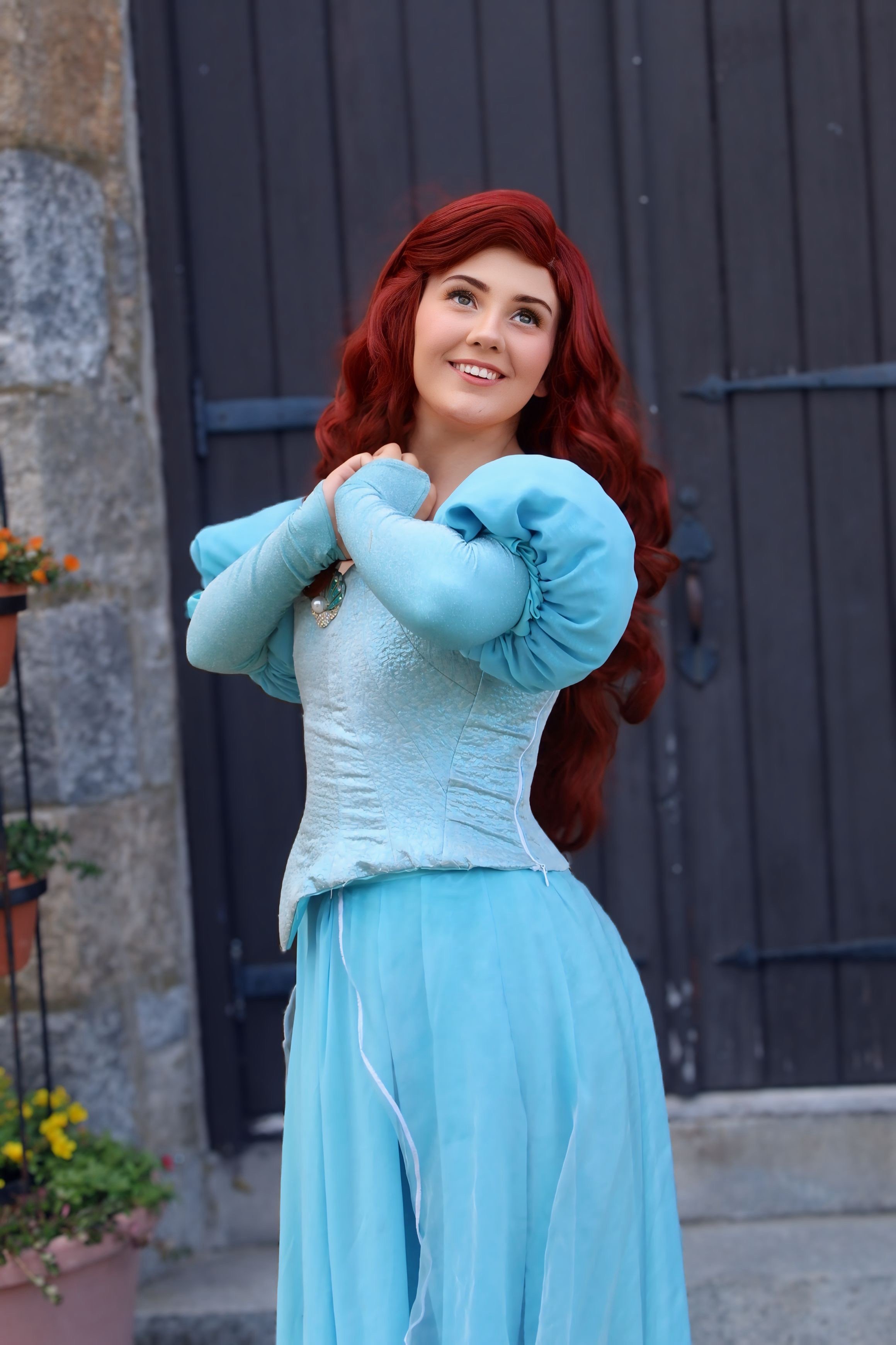 A woman dressed as Ariel from The Little Mermaid, with long red hair and a blue dress, standing in front of green foliage.