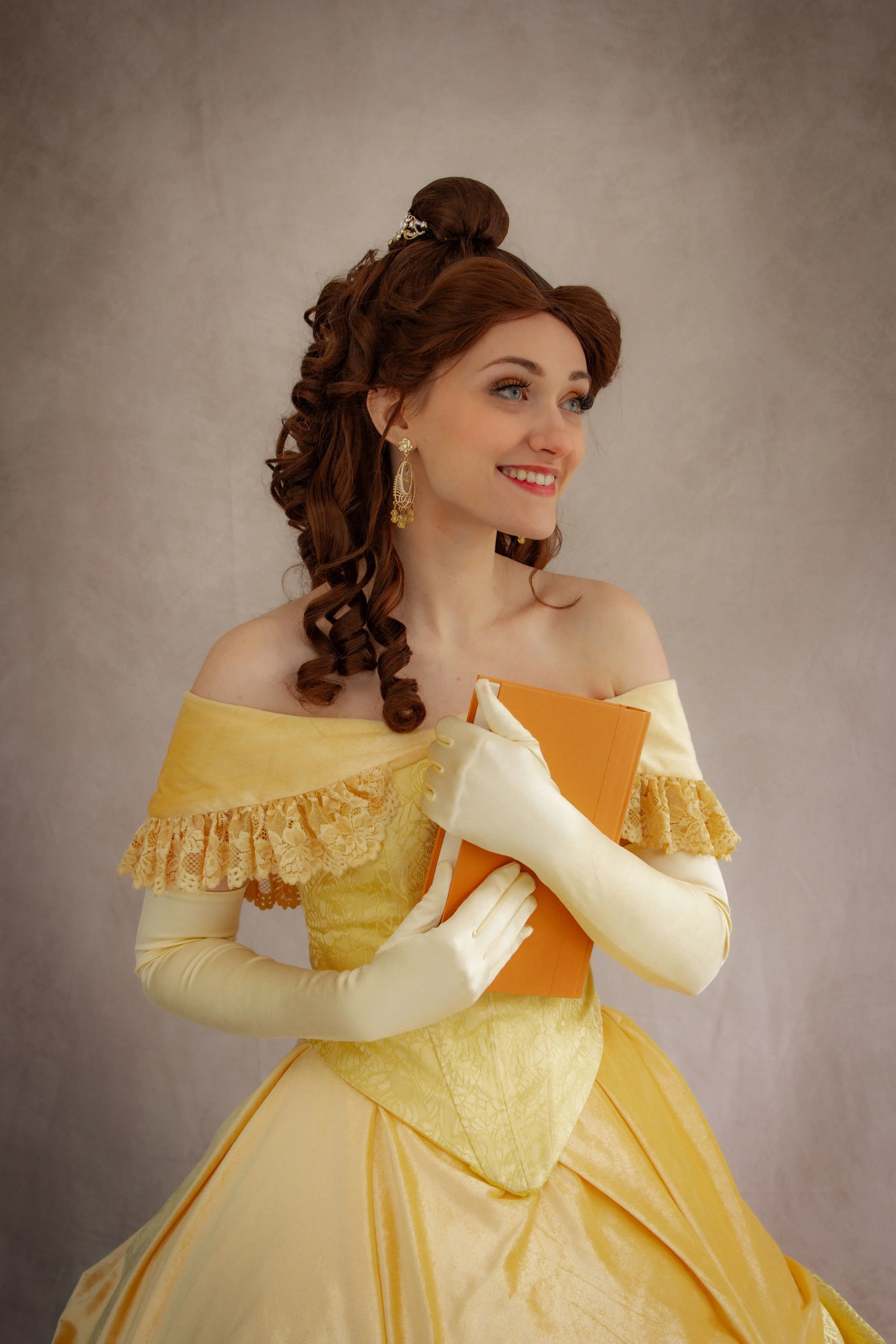 A woman dressed as Belle from Beauty and the Beast, smiling, wearing a yellow gown with gloves, with pink and pastel-colored paper butterflies and balloons in the background.