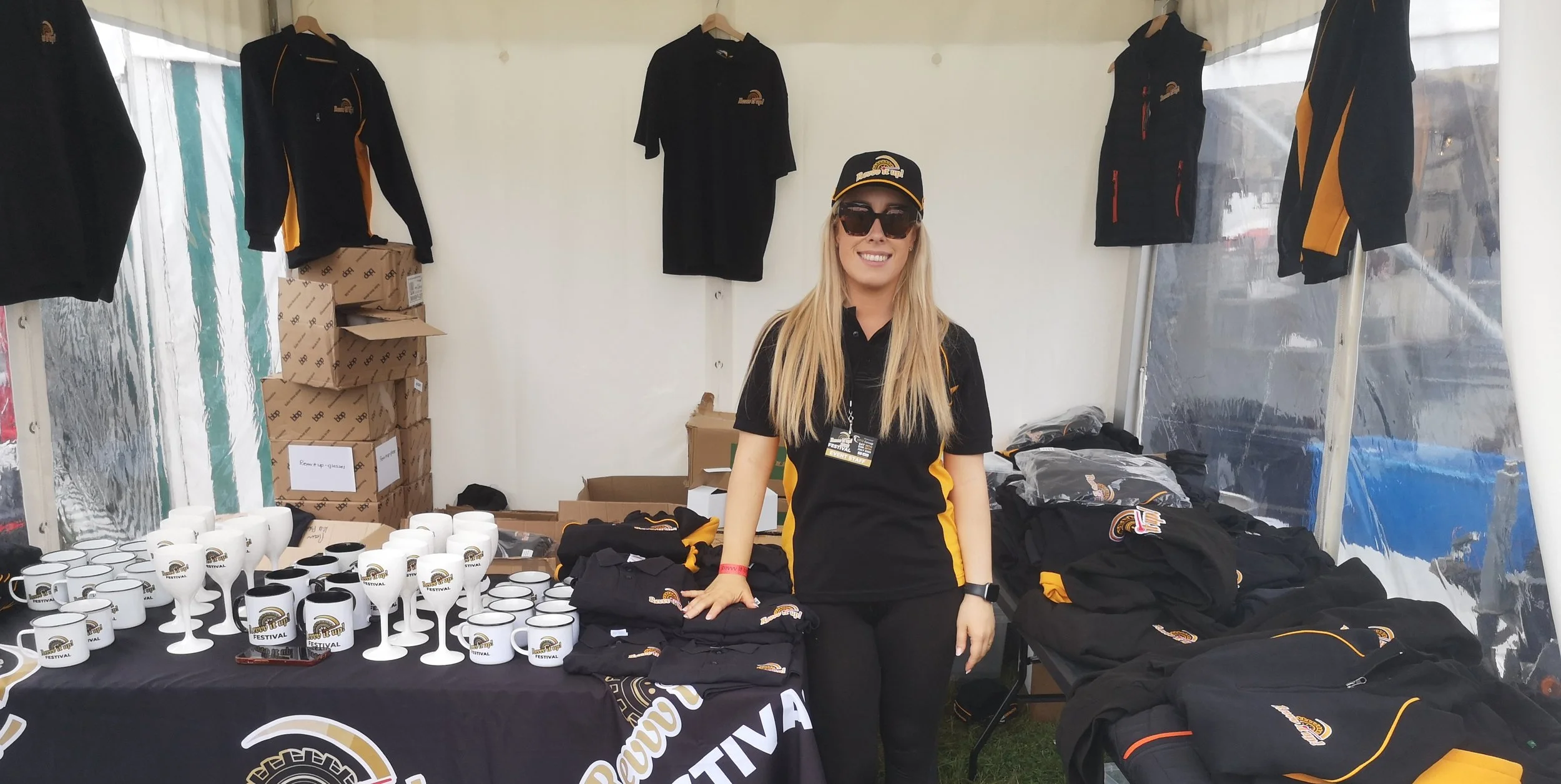 A woman standing in a booth selling team merchandise, including mugs and clothing with a racing emblem, at an outdoor event.