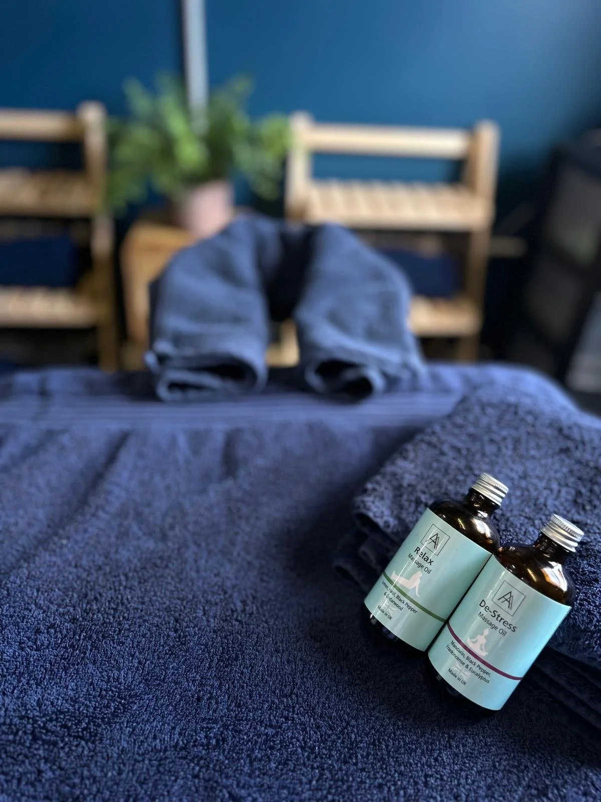 Two bottles of massage oil labeled 'Relax' and 'De-Stress' on a massage table with rolled towels. In the background are two wooden benches with plants behind them.