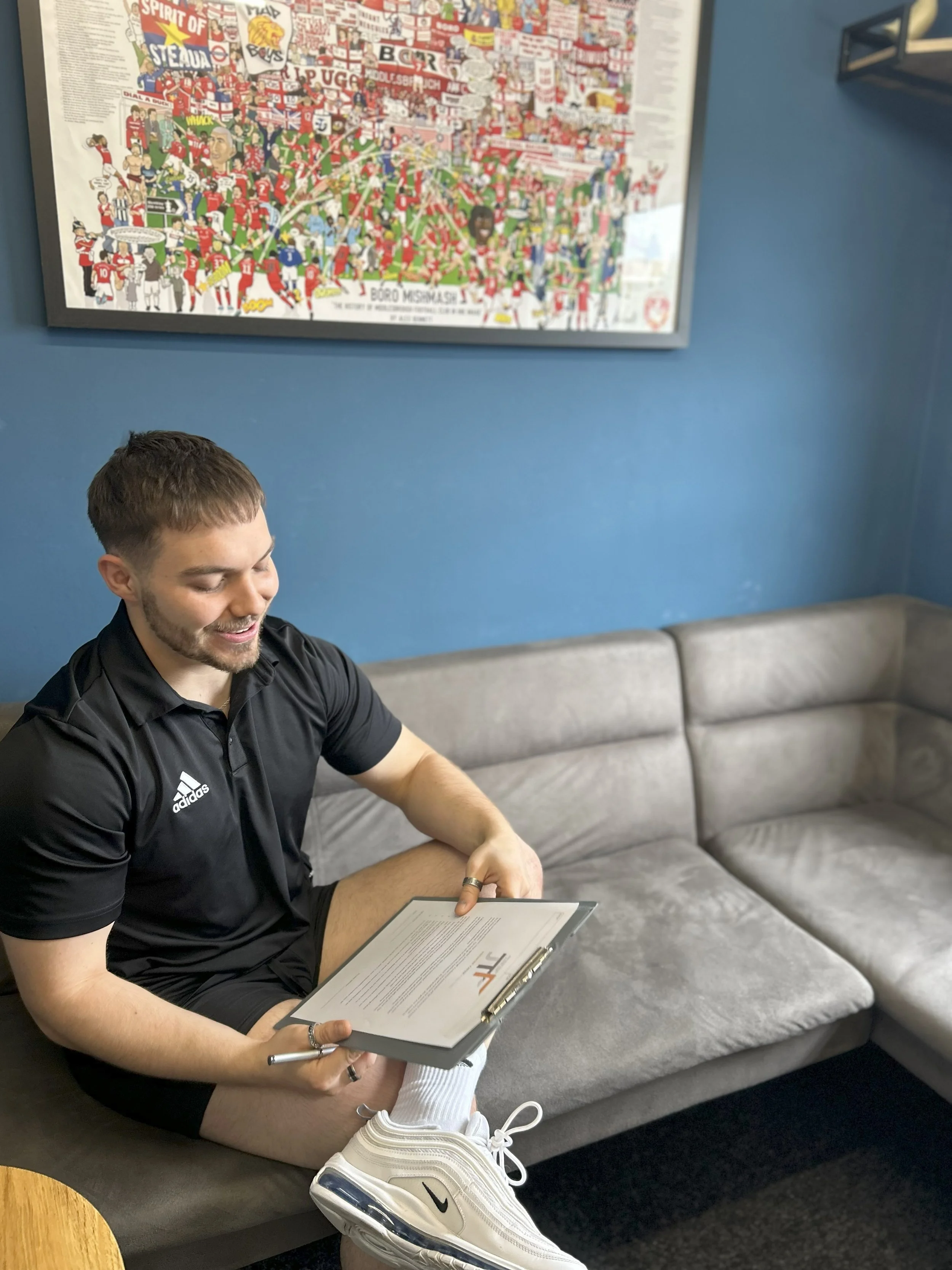 A young man sitting on a gray couch, smiling, while holding a clipboard and a pen. He is wearing a black Adidas sports shirt, black shorts, white Nike sneakers, and white socks. Behind him, there is a blue wall with a large, colorful illustrated poster.