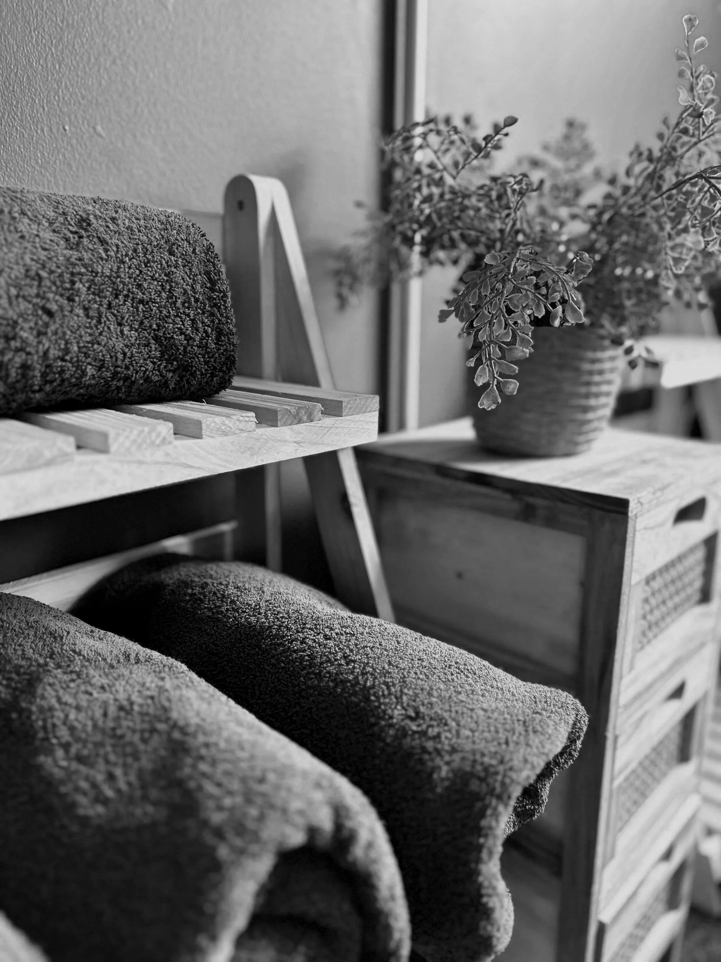 Black and white photo of cozy textured blankets on a wooden bench and a basket of plants on a wooden side table.