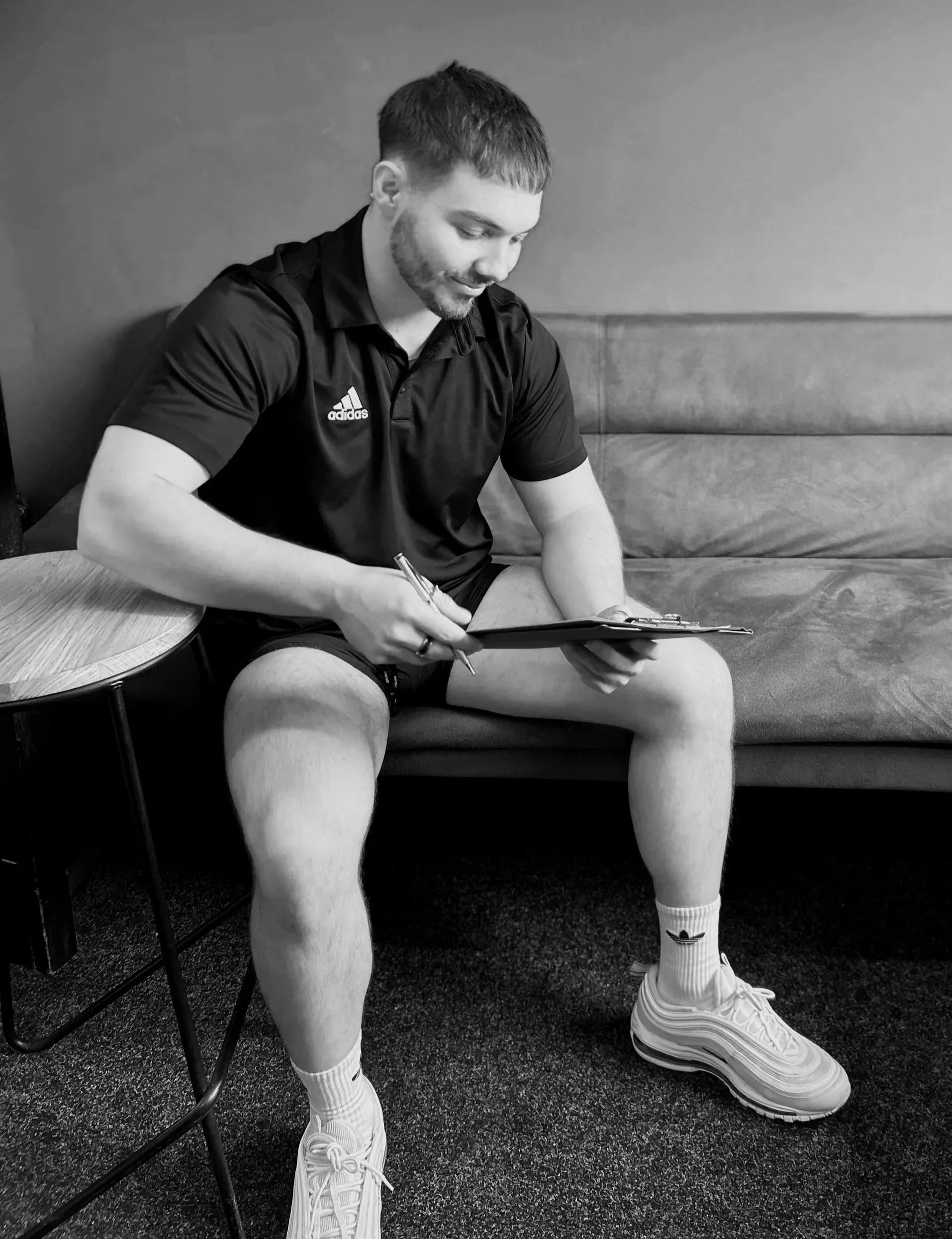 A man sitting on a couch with a clipboard and pen, wearing a sports shirt, shorts, socks, and sneakers, in a room with a table and carpeted floor.