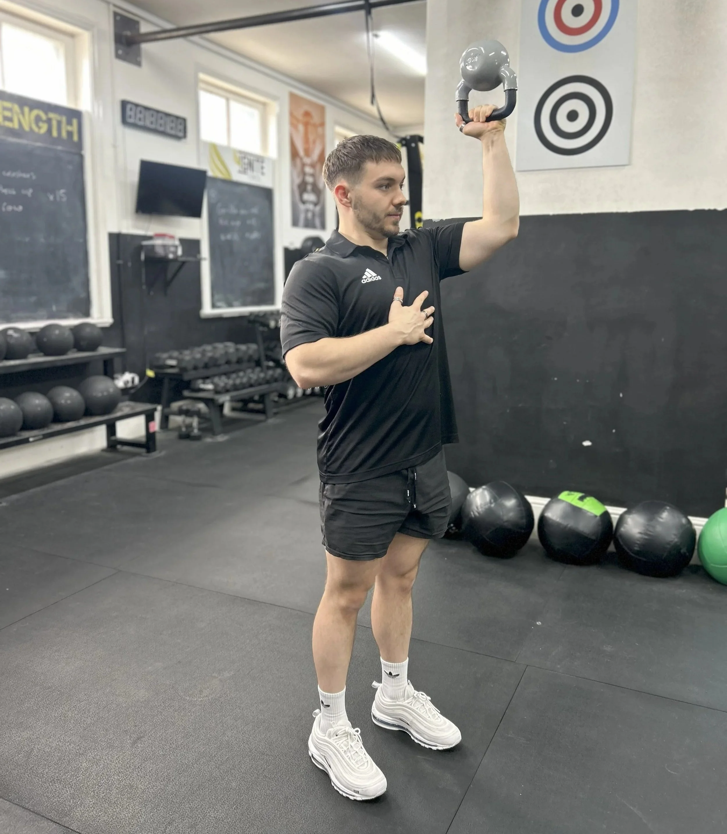 A man in black athletic attire performs a kettlebell exercise in a gym, holding the kettlebell overhead with one hand and placing his other hand on his chest.