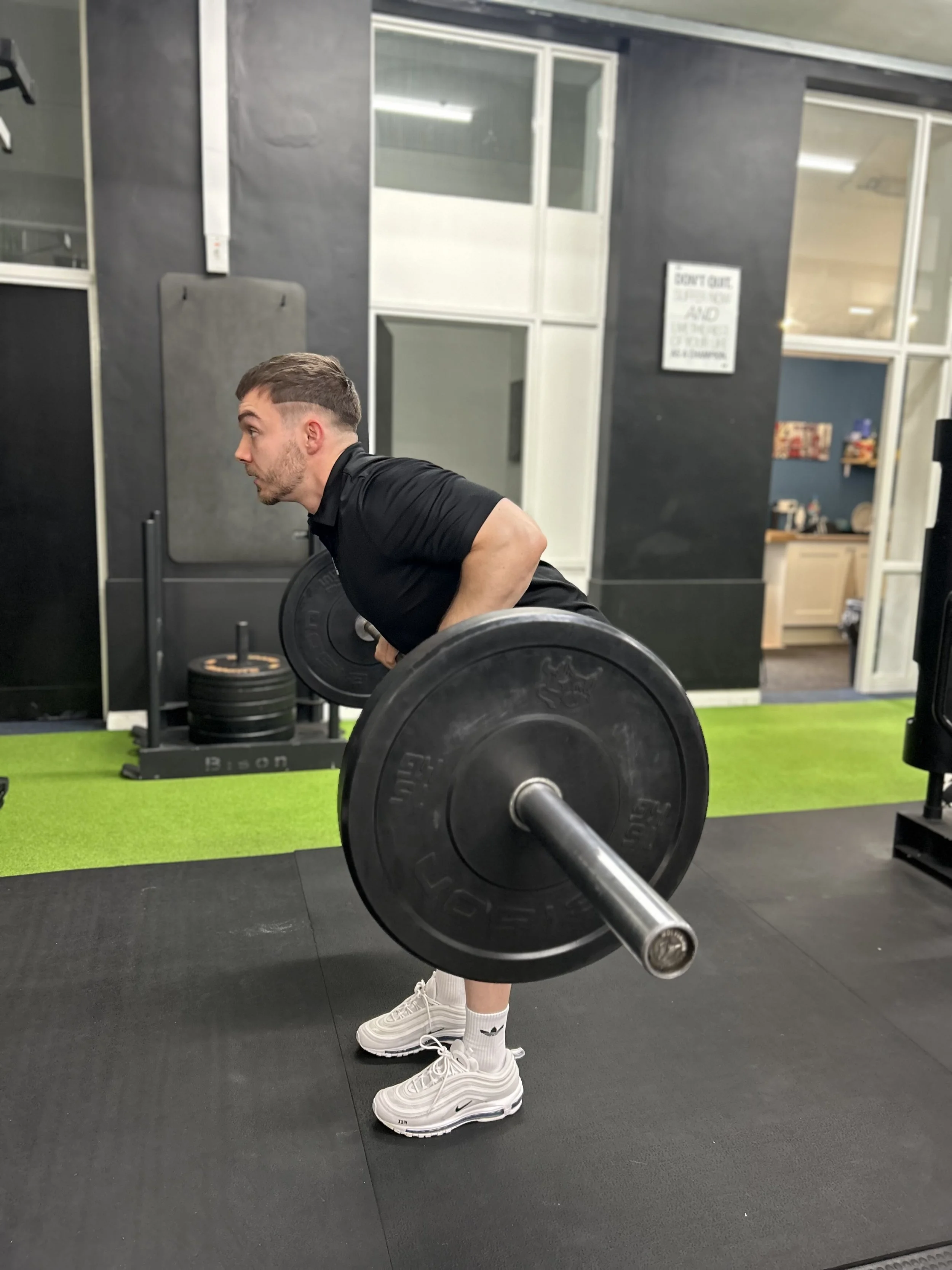 A man in a black shirt and white sneakers lifting a barbell with weights at a gym. He is bending forward with his back straight, in the middle of a workout.