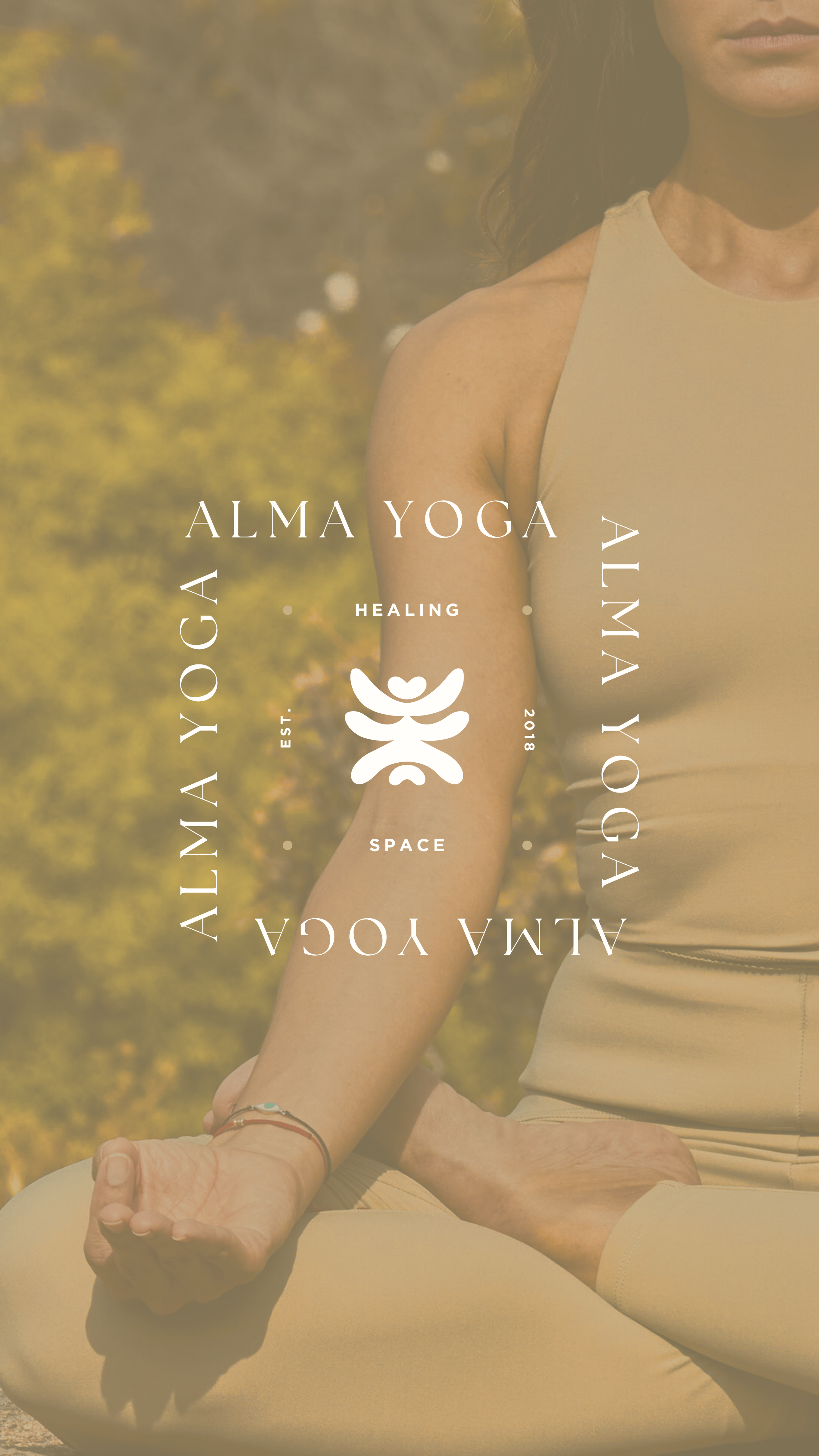 A woman in a beige athletic tank top and leggings practicing yoga outdoors in a cross-legged pose with her hand resting on her knee, surrounded by nature with autumn trees in the background.