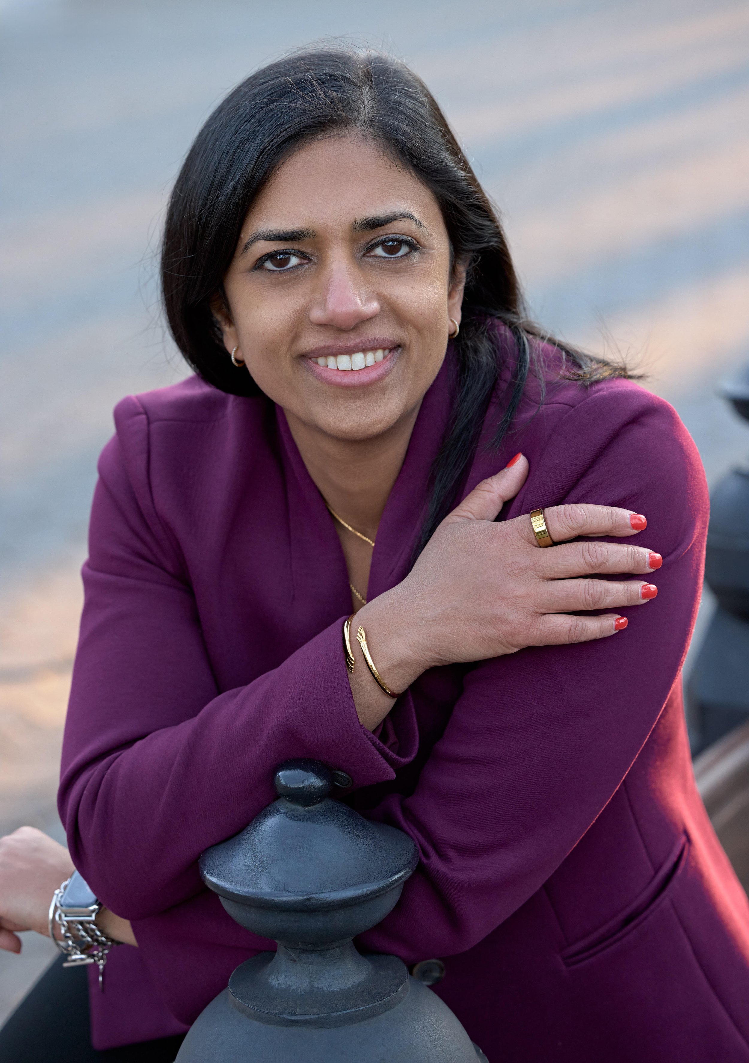 Portrait of Vasanta Pundarika, founder of Lotuspring, with shoulder-length black hair, wearing a purple jacket, gold jewelry, and red nail polish, sitting outdoors with a cloudy sky background.