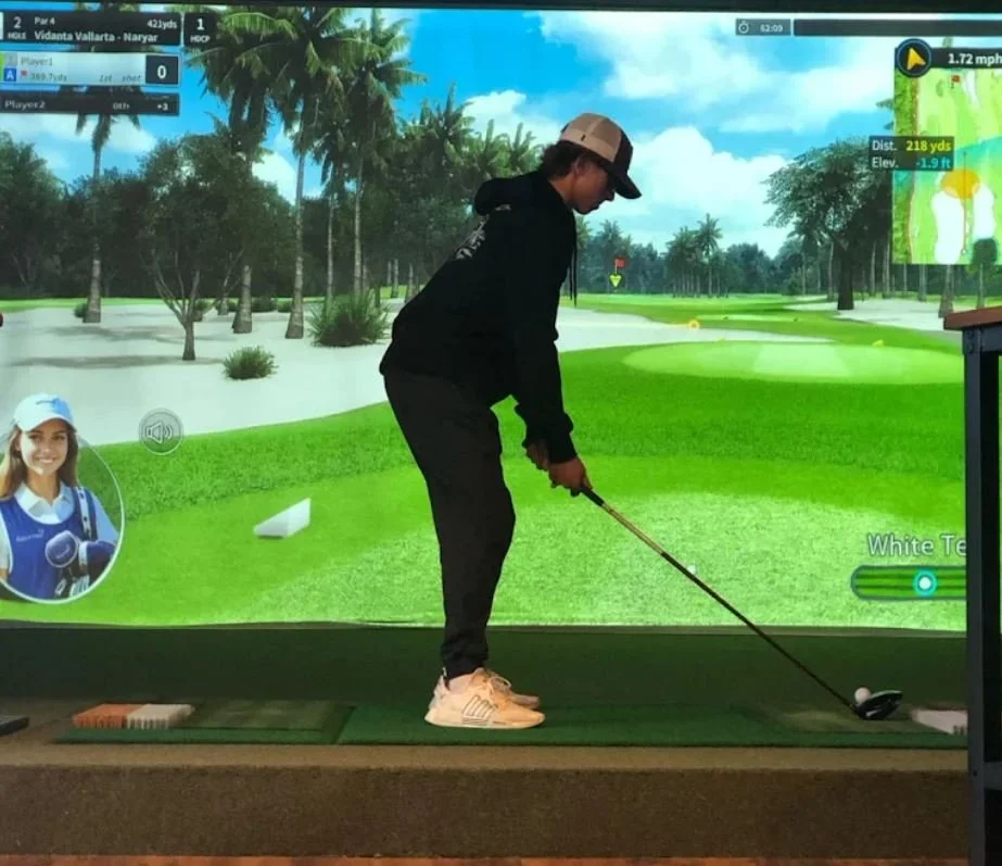 A person playing golf on an indoor simulator, with a virtual golf course on the screen. The person is standing at the tee, holding a golf club, preparing to hit the ball. The screen shows a tropical landscape with palm trees and blue skies.