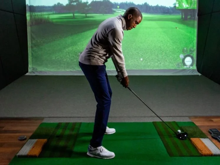 Man practicing golf swing in an indoor golf simulator with a virtual golf course projected on the screen behind him.