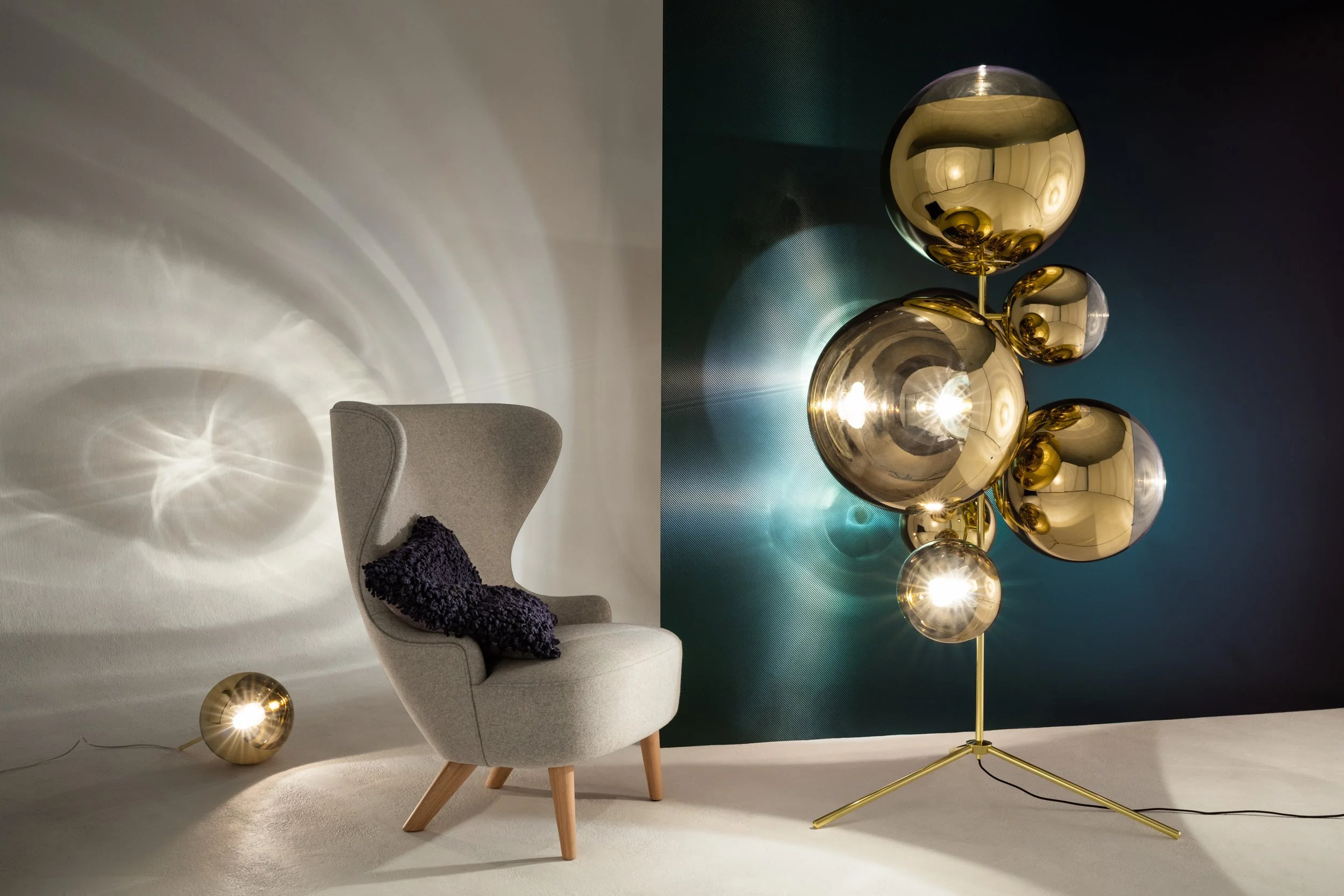 A modern gray armchair with a dark purple textured pillow, two spherical gold-colored lamps on the floor and a large multi-globe gold and glass chandelier.