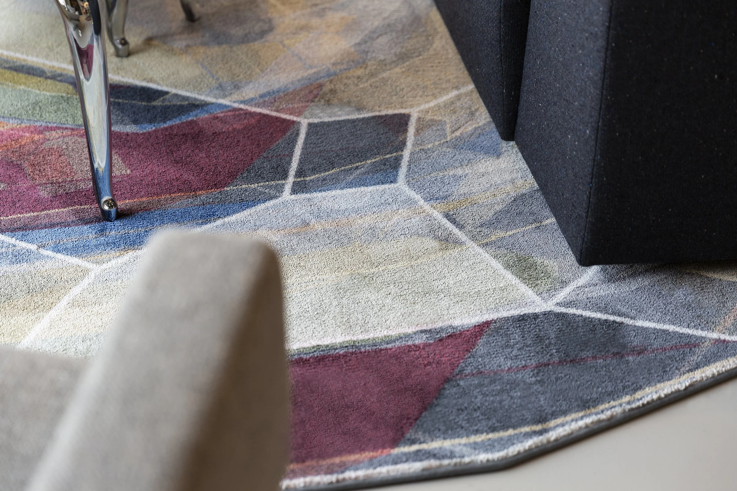 Close-up of a colorful patterned Moooi Carpets rug beneath a beige armchair, a black sofa, and part of a metallic chair leg.