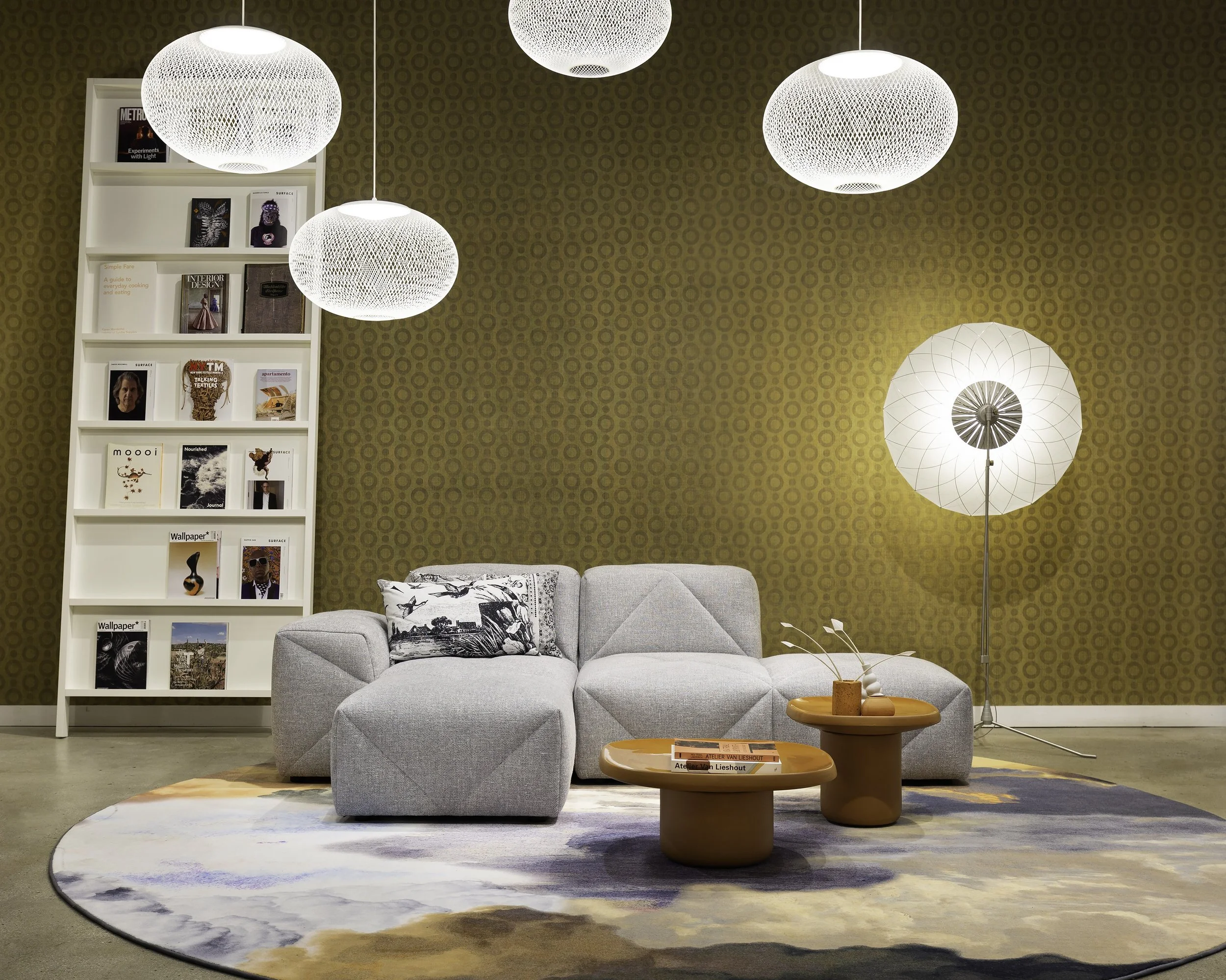 Living room with gray sectional sofa, round coffee tables, white bookshelf with books, contemporary floor lamp, and paper lantern hanging lights, on a colorful abstract rug.