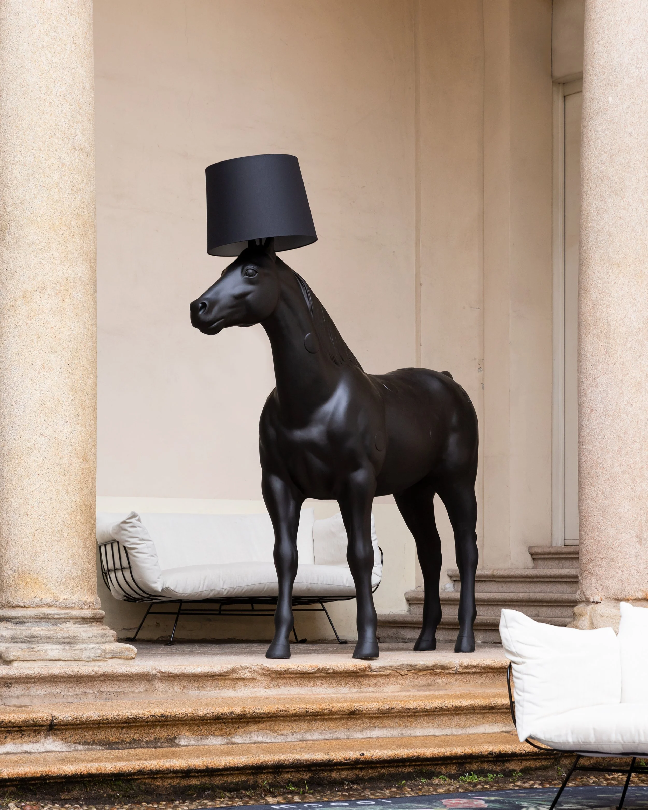 Black horse-shaped lamp from Moooi with a lampshade on its head, sitting on stone steps near an outdoor patio with white pillows and seating.