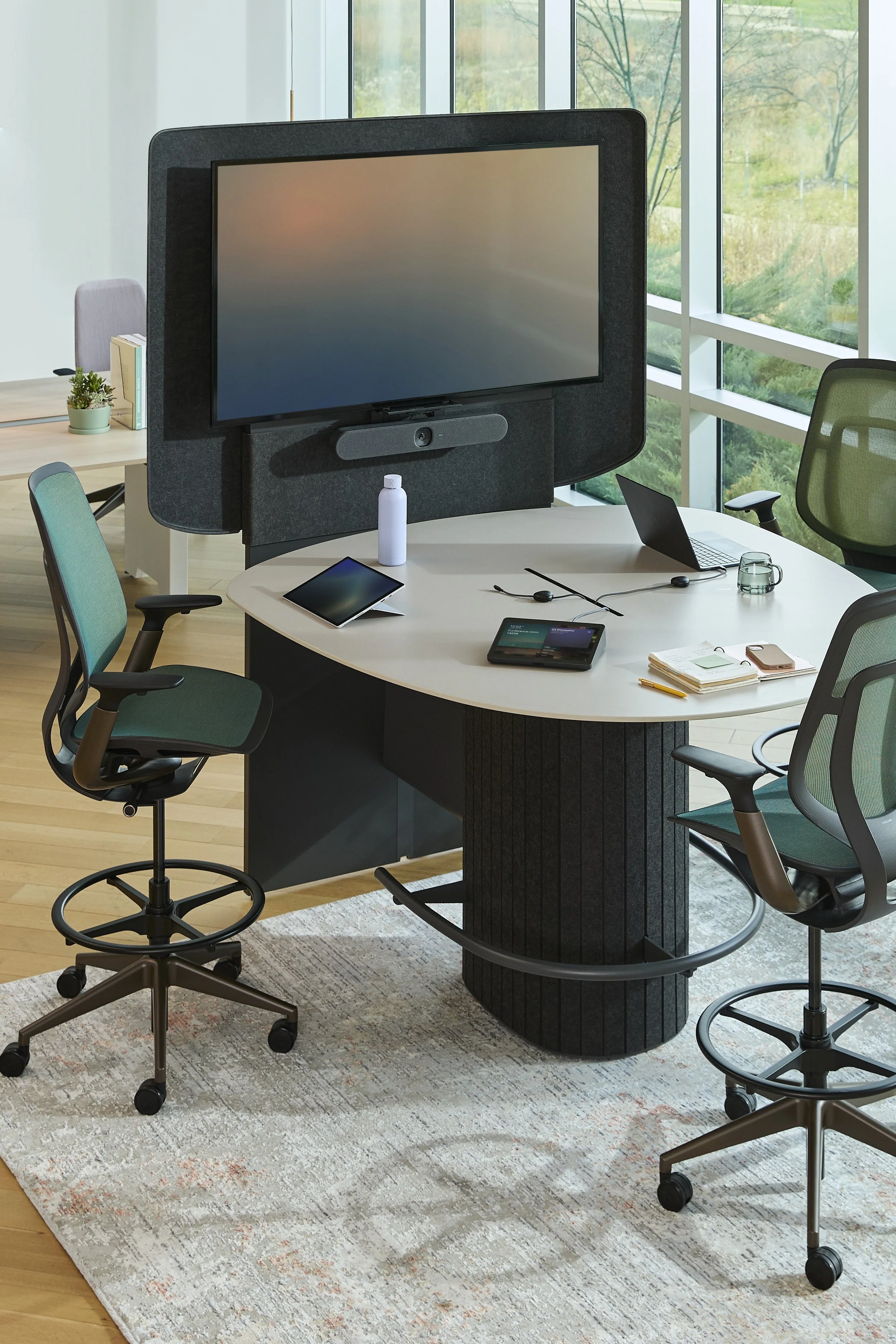 Modern conference room with a large oval table, chairs, and a wall-mounted screen.